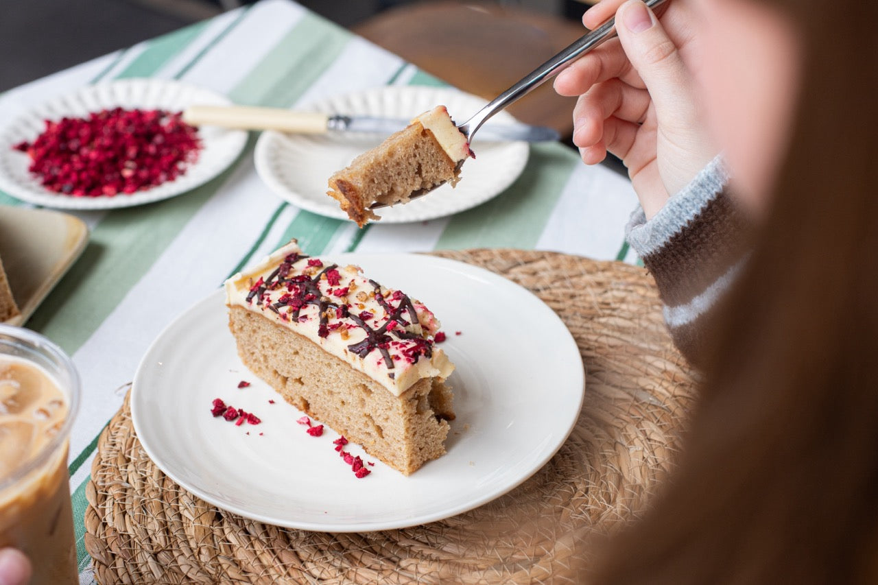 Person cutting into a slice of Banana Split cake with a fork on a table with a glass of iced coffee and a plate of pomegranate seeds.
