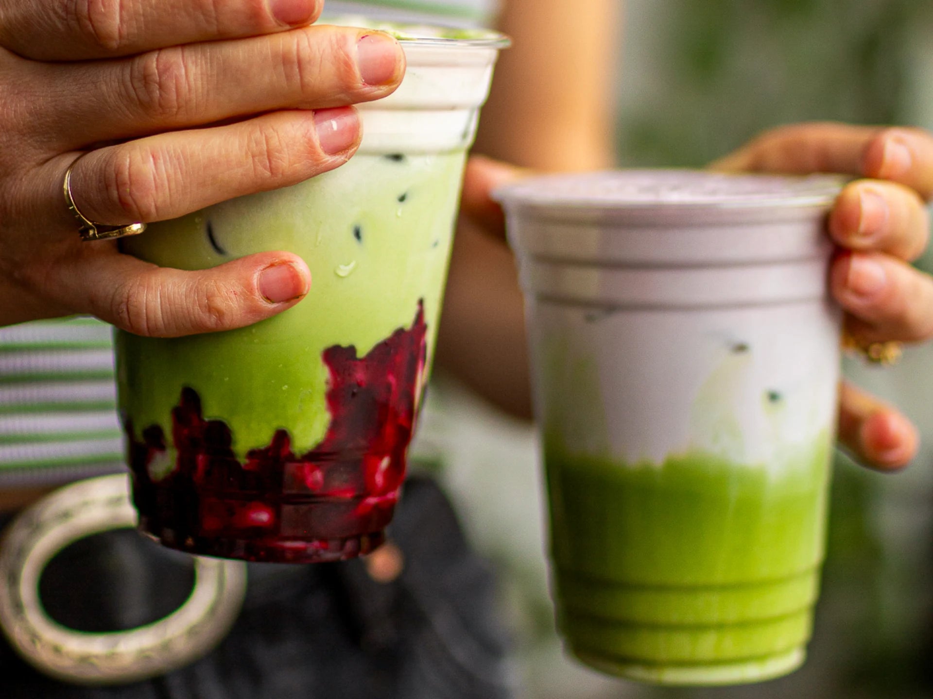 Two hands holding colorful Ceremonial-Quality Wakaba Matcha drinks in plastic cups against a blurred background