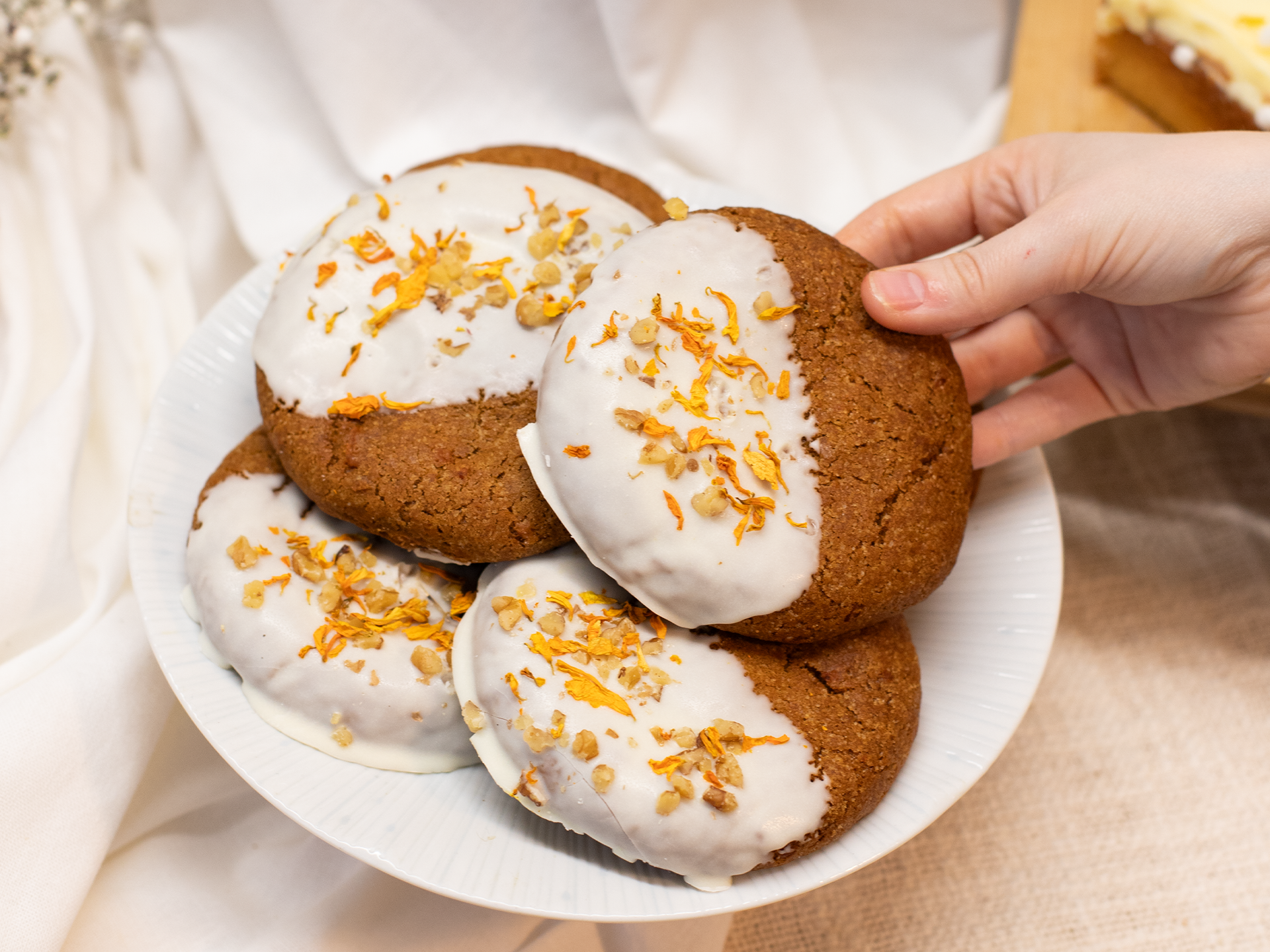 Hand holding a plate of Carrot Cake Cookie with white icing and orange sprinkles.