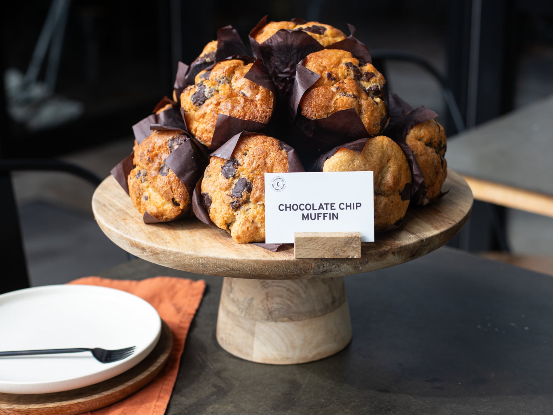 Wooden cake stand with chocolate chip muffins and a sign indicating the type of muffin.