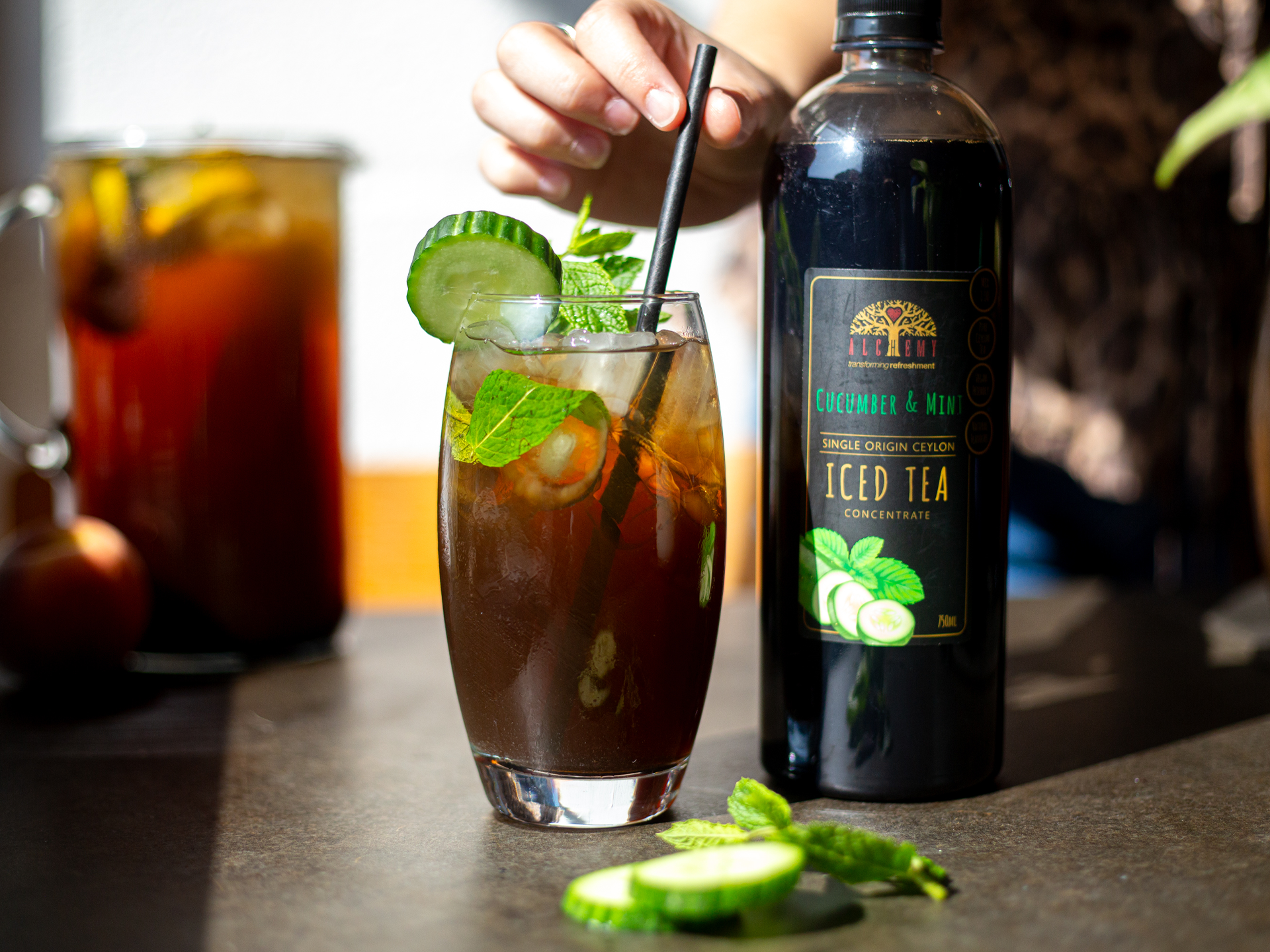 Glass of iced Alchemy tea with garnishes next to a bottle of iced tea on a table.