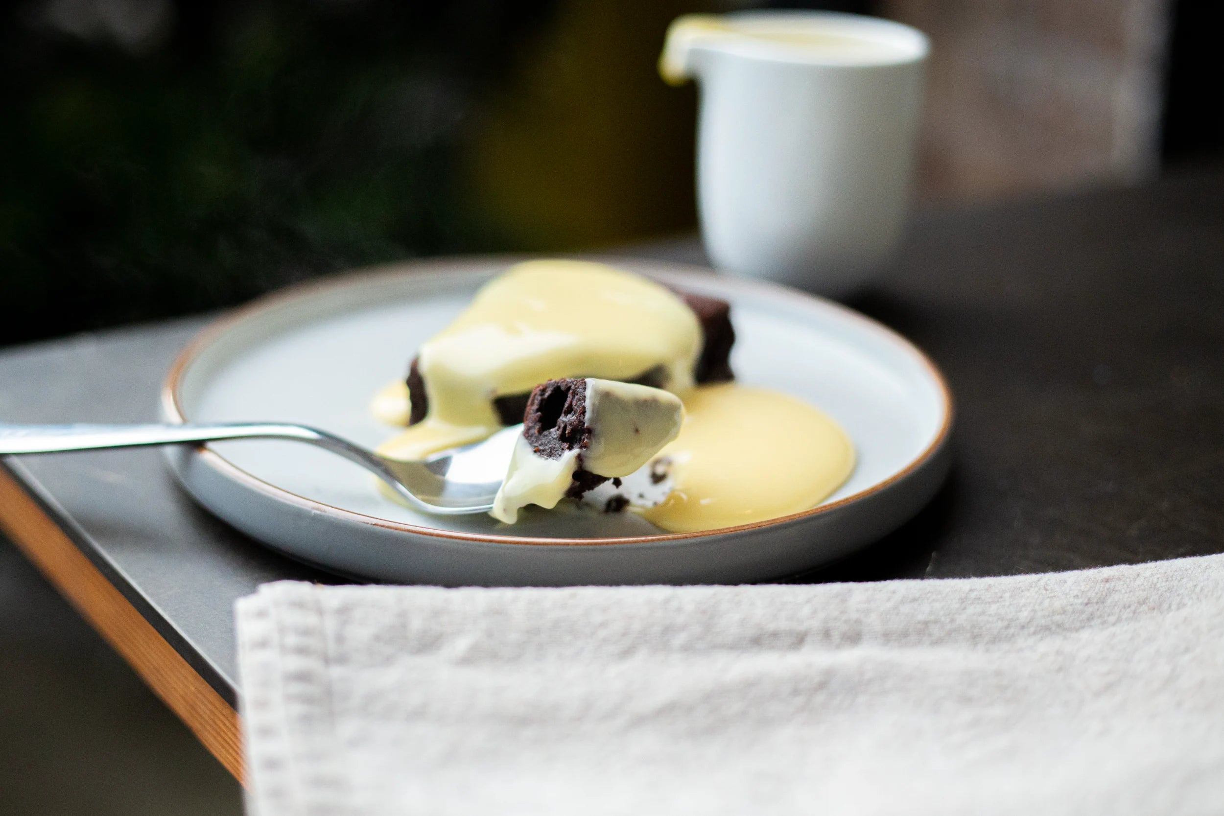 Dessert Brownie with a side of custard on a plate, with a fork and a small pitcher in the background.