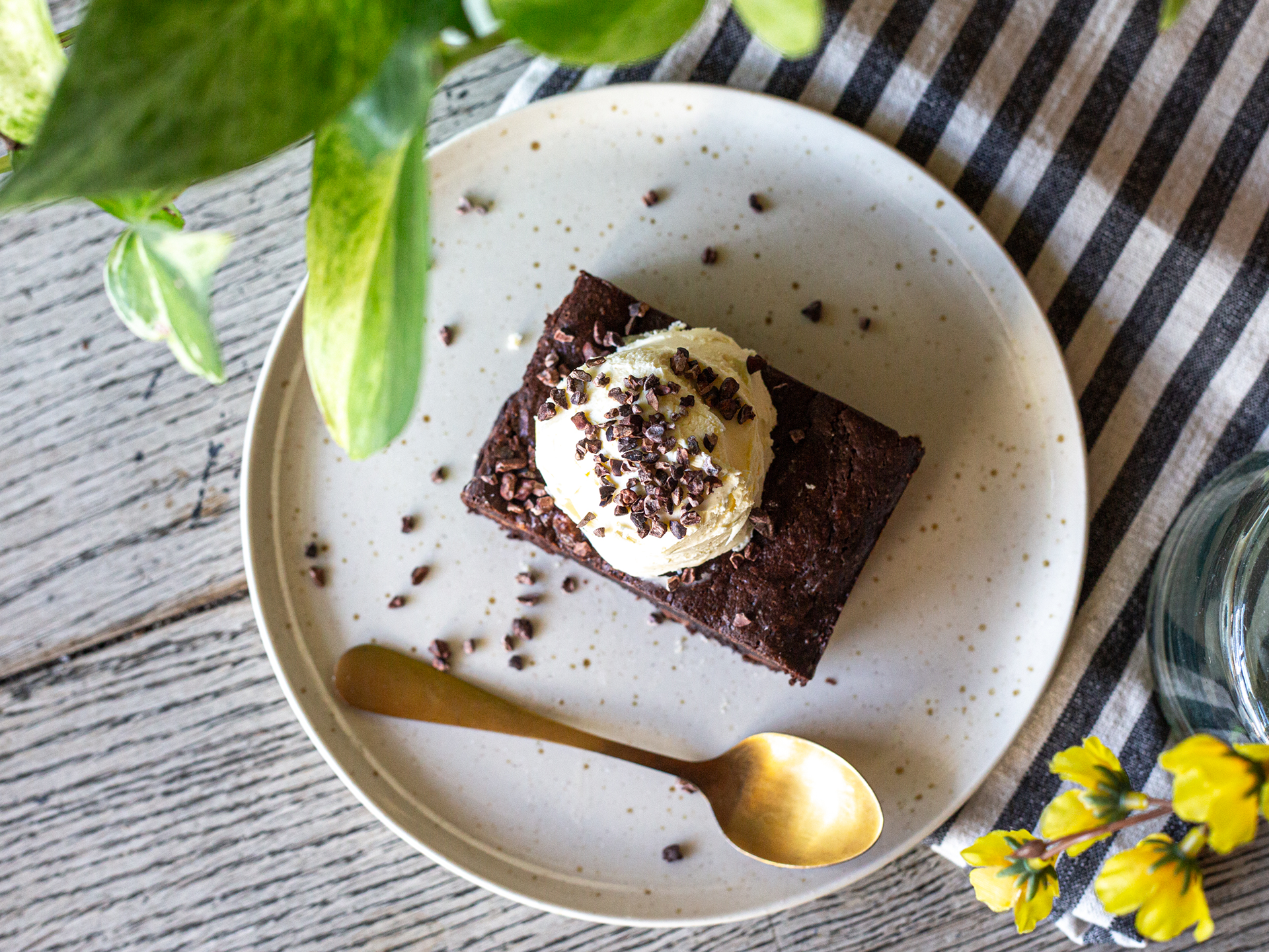 Brownie with ice cream on a speckled plate with a gold spoon, on a wooden surface with a striped cloth and plant.