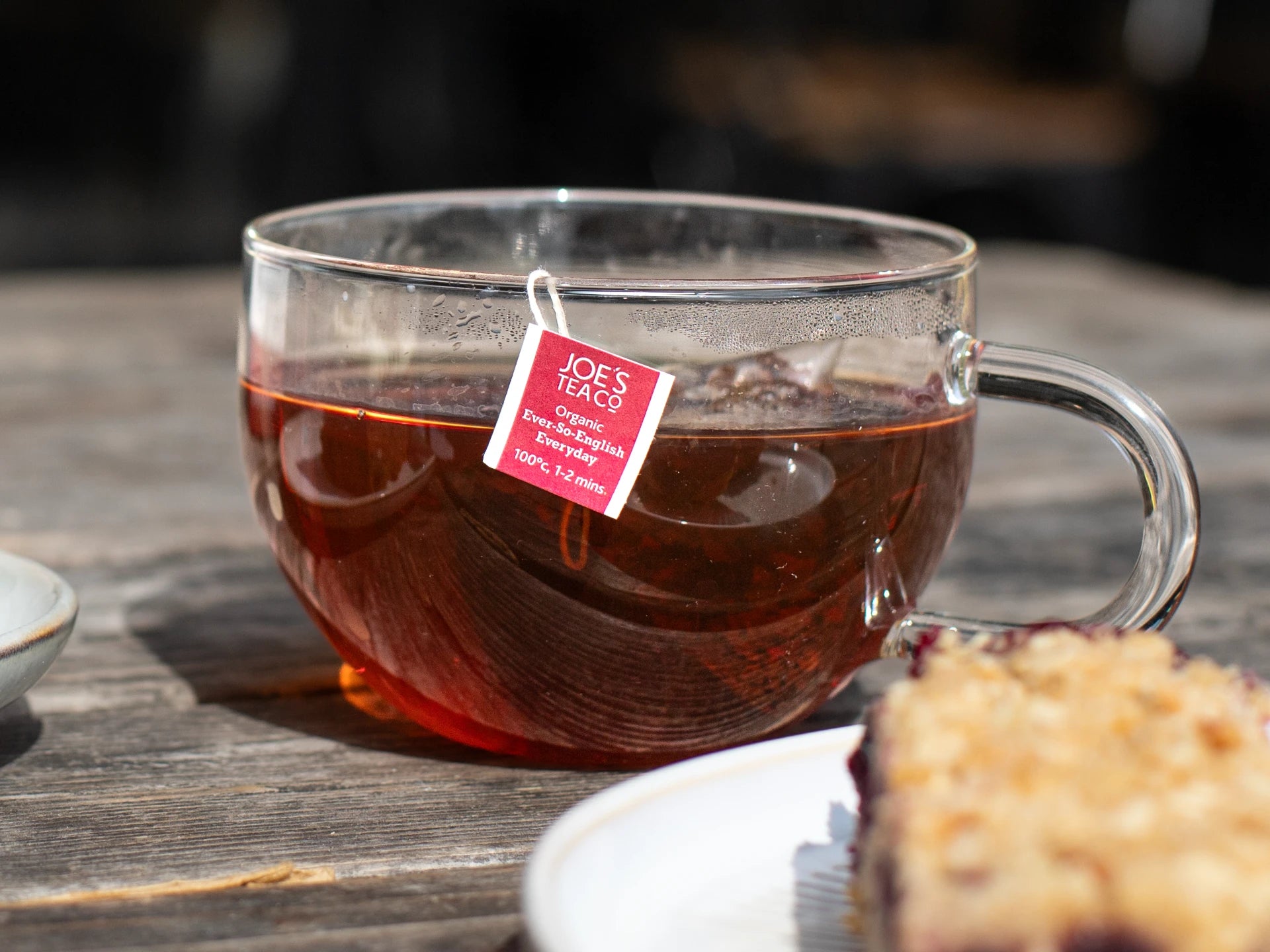 Glass mug of Joe's Tea Co - Ever-So-English Breakfast tea with a visible tea bag on a wooden surface.