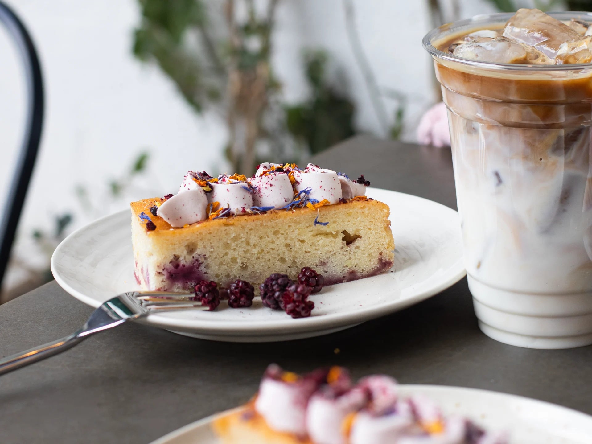 Slice of Lemon Blackberry Elderflower cake with berries on a plate next to a glass of iced coffee.
