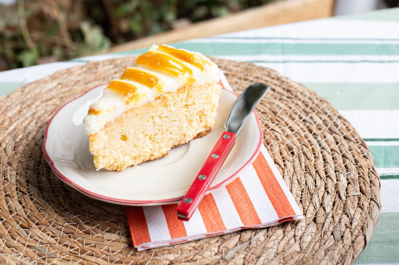 Mango Pineapple Coconut cake Slice of cake with white frosting and orange slices on a plate with a red knife, on a woven mat.