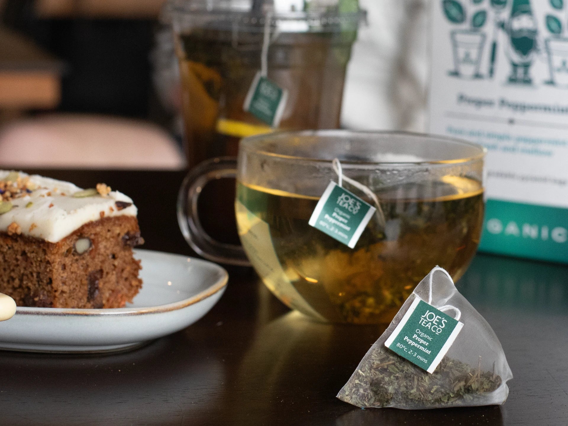 Joe's Tea Co - Proper Peppermint Tea cup with a tea bag, cake on a plate, and a box of tea on a table.
