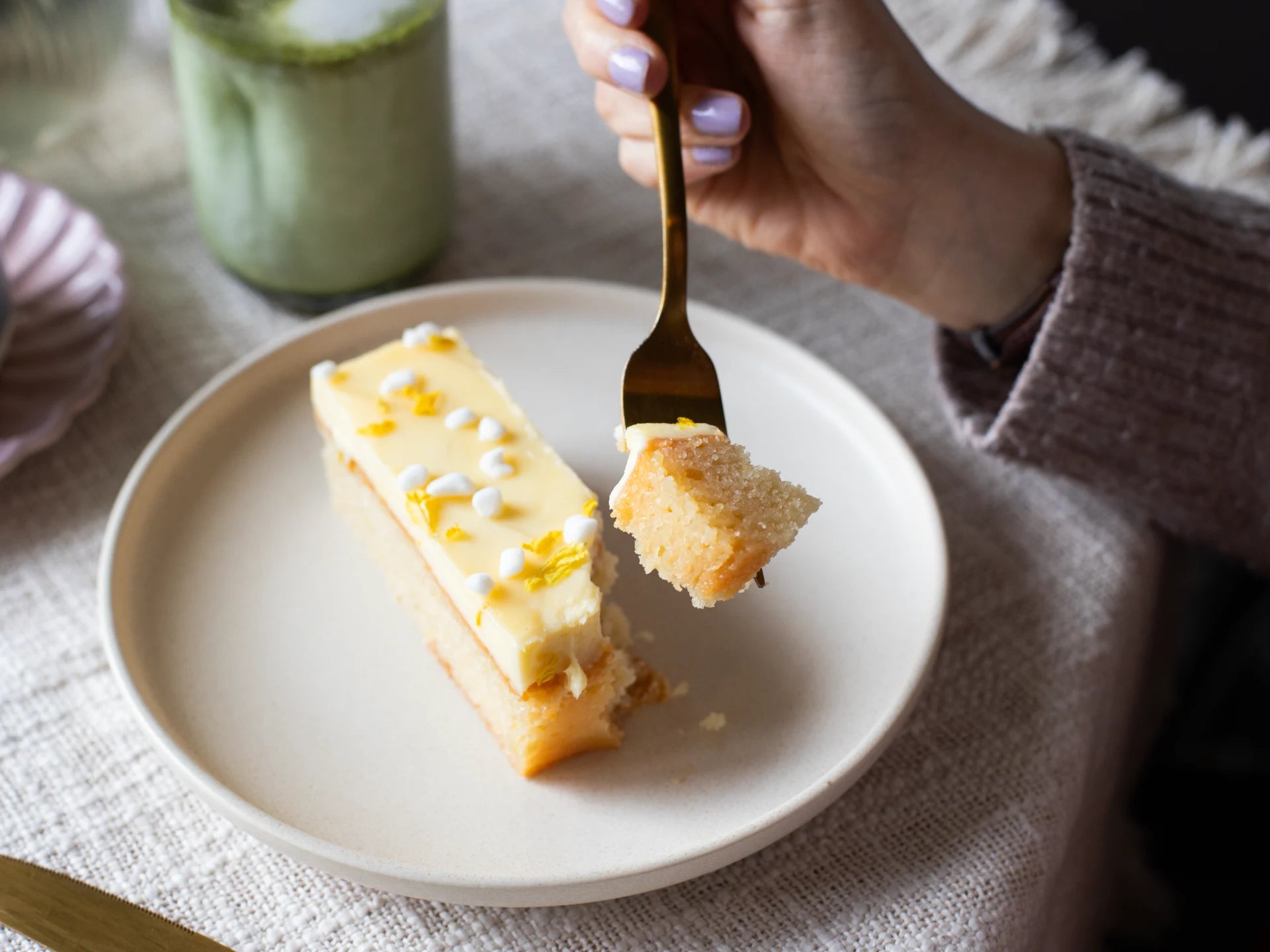 Person eating a piece of Raspberry Rhubarb Cake with a fork on a white plate, with a green cup in the background.