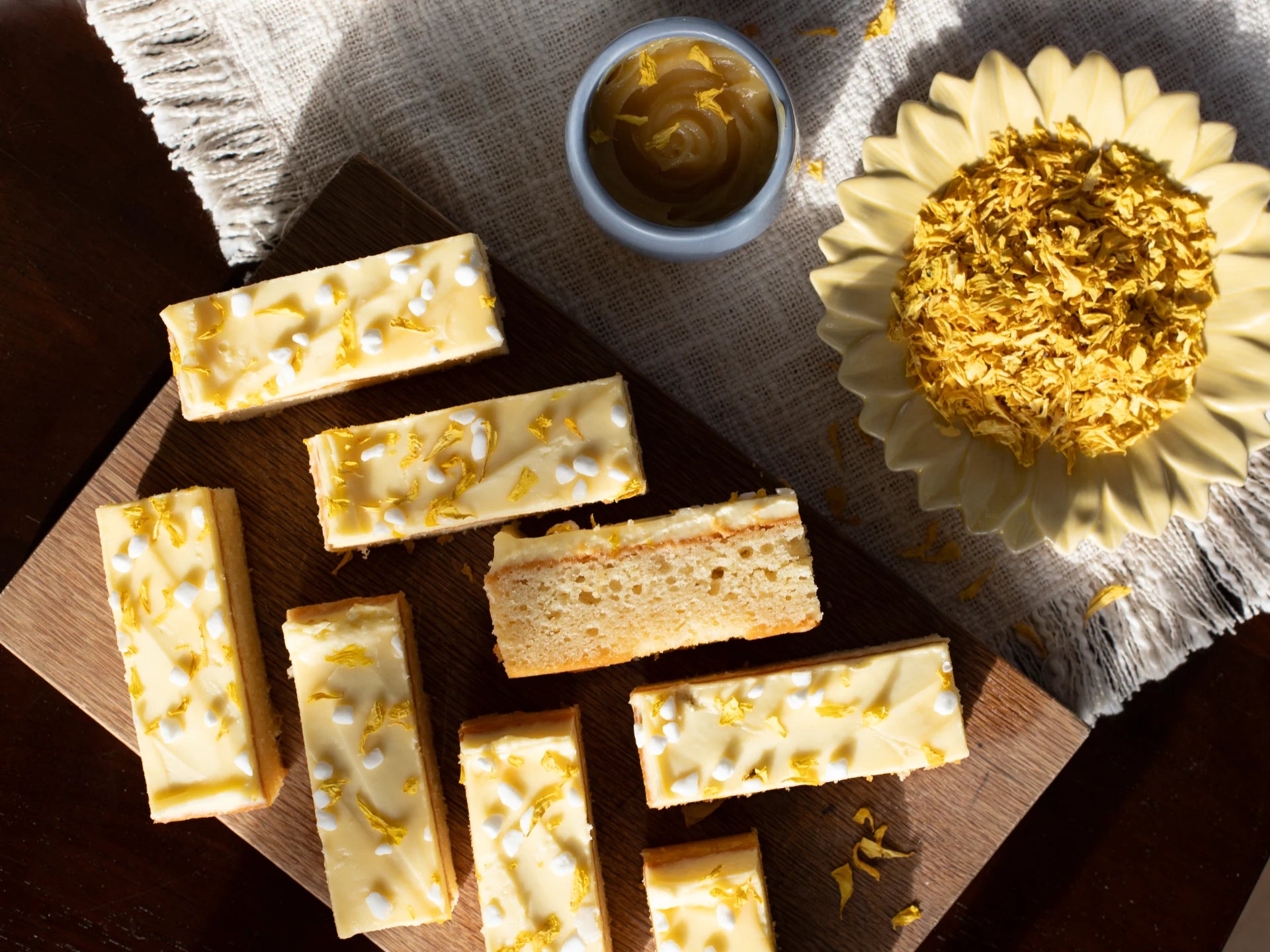 Sicilian Lemon Meringue with a white coating and gold sprinkles on a wooden board, with a small bowl of gold powder and a sunflower in the background.