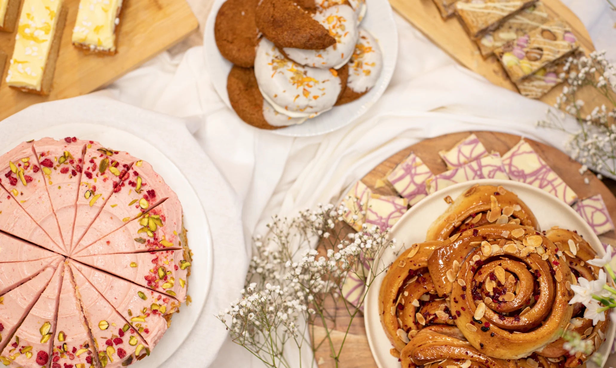 Assorted spring cakes including a pink cake, cinnamon rolls, and cookies on a white surface.