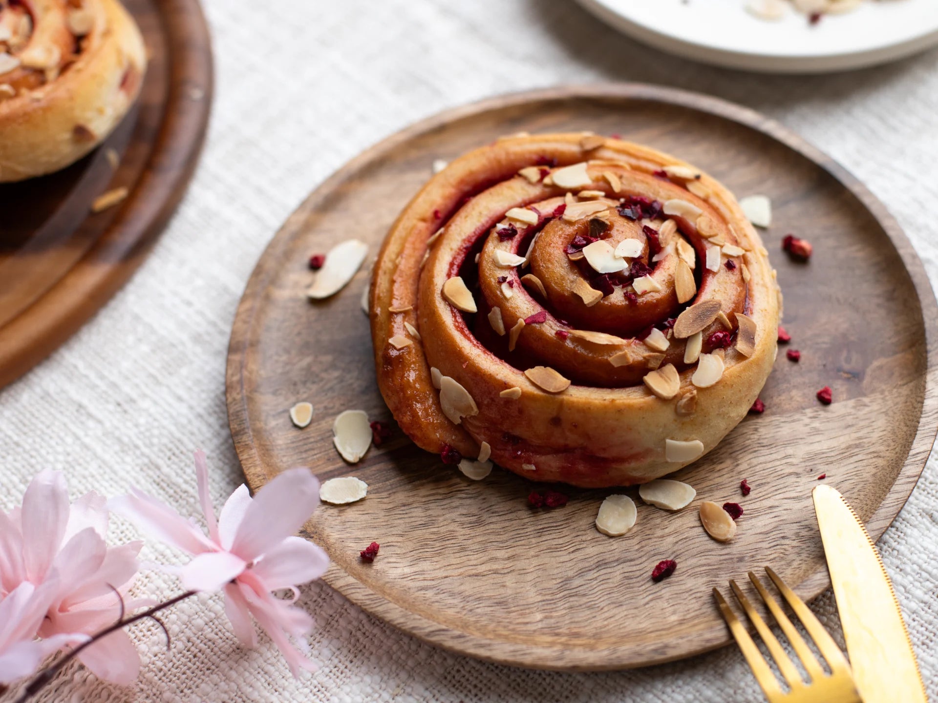Sticky Cherry Bakewell Bun with almonds on a wooden plate with a gold fork and pink flowers.
