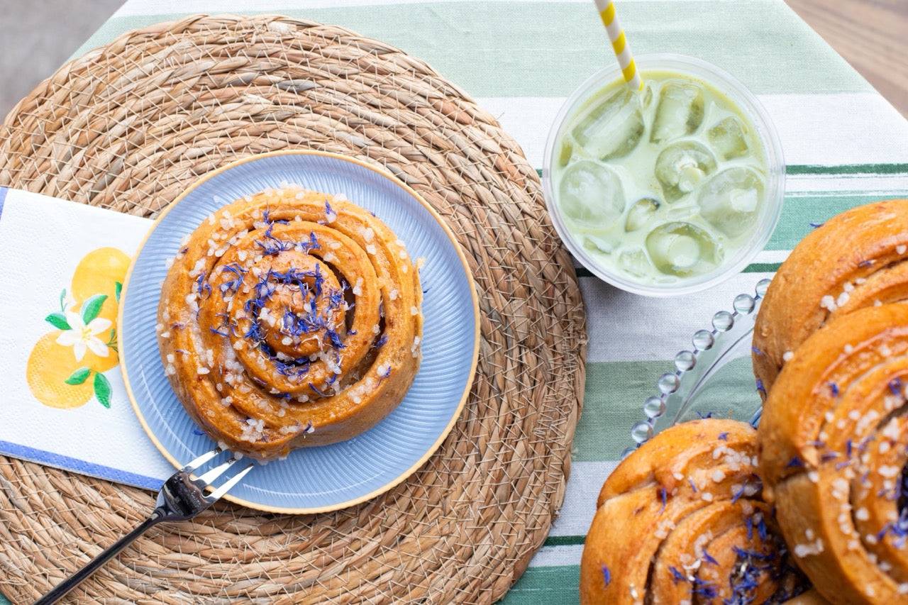 Sticky Lemon Drizzle buns with purple sprinkles on a blue plate, surrounded by other cinnamon rolls and a drink with a straw on a woven placemat.