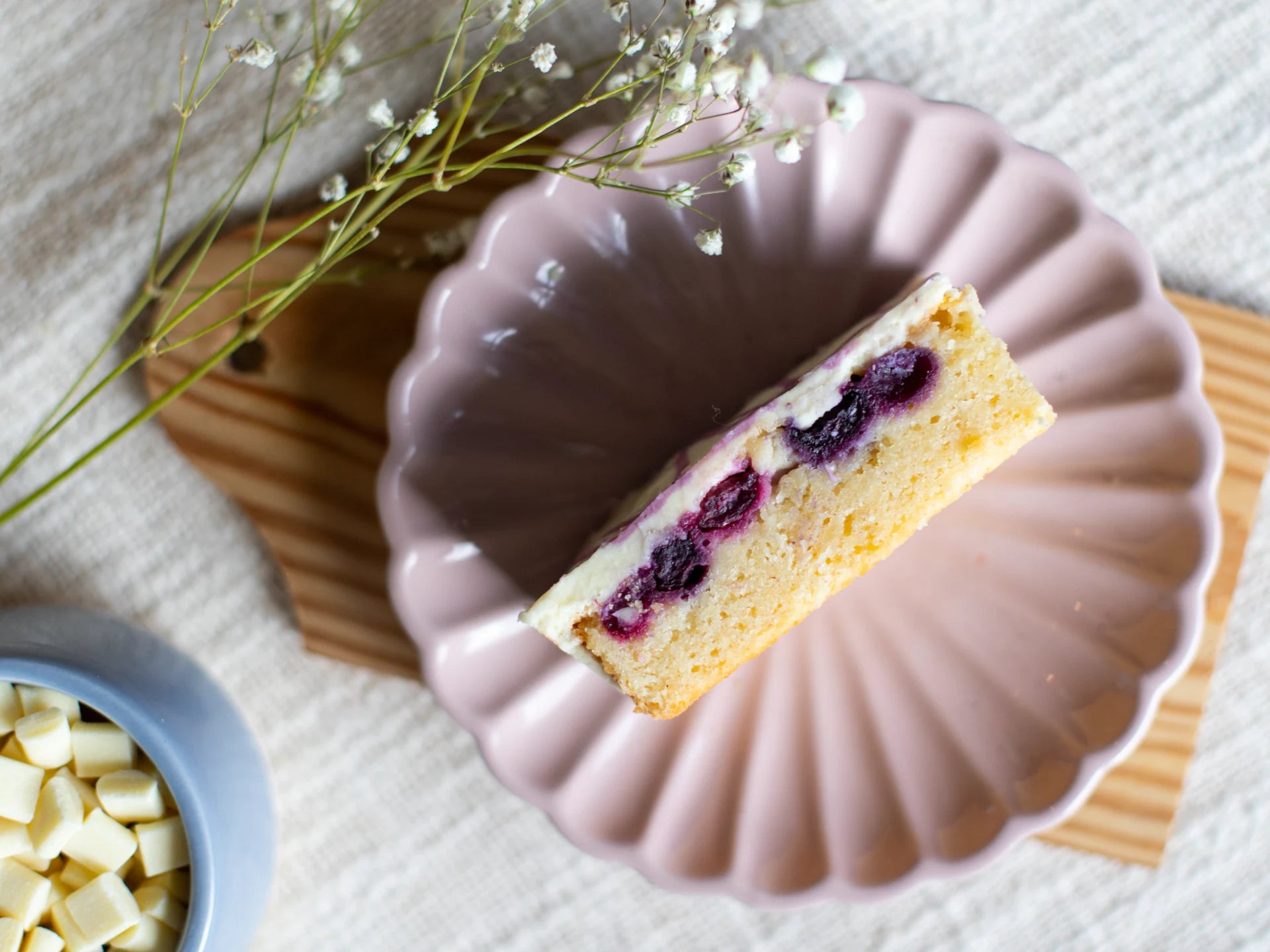 Slice of White Chocolate Blueberry Blondie cake with berries on a pink cake stand, surrounded by white flowers and a bowl of white chocolate pieces.