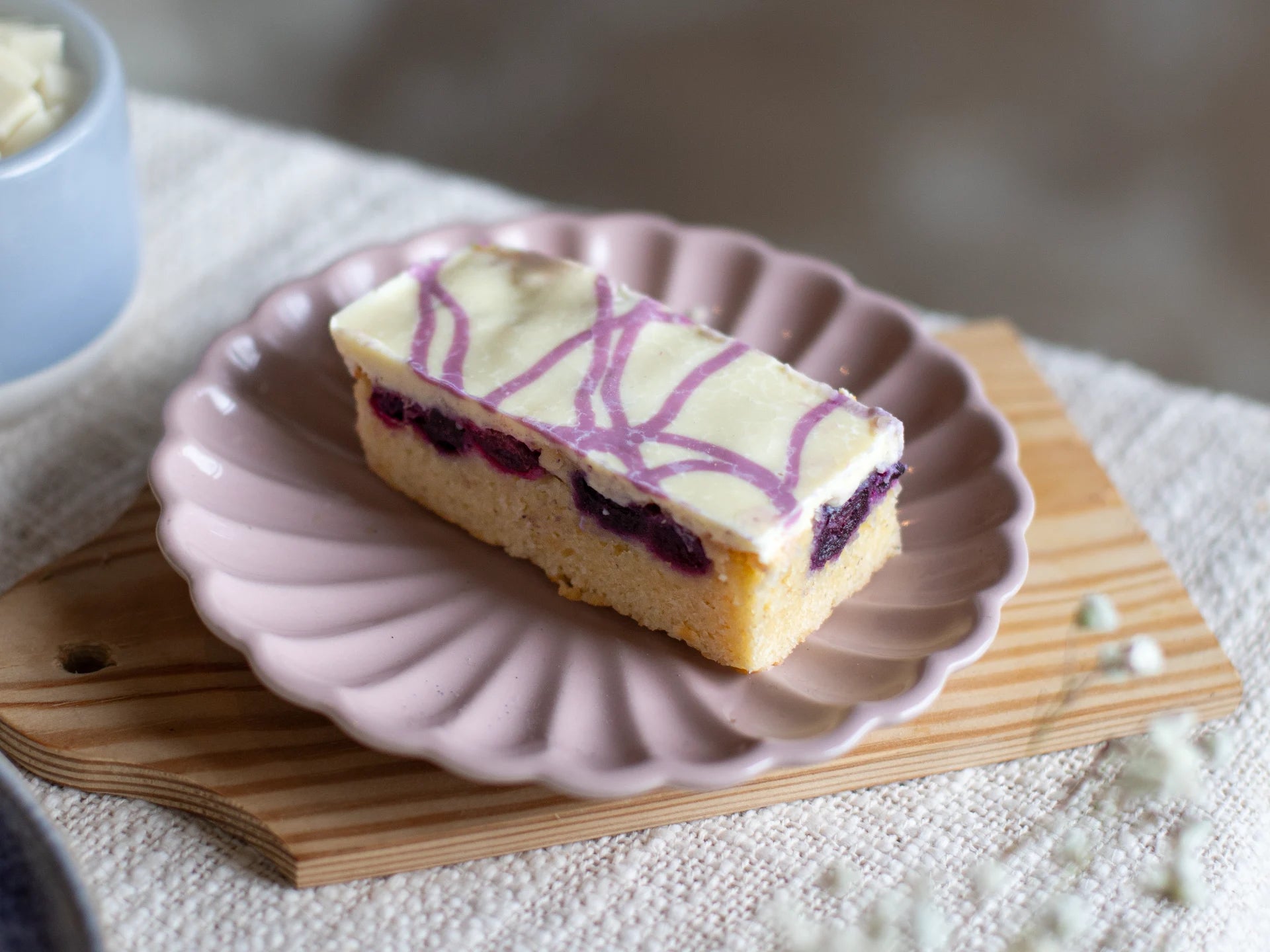 Rectangular White Chocolate Blueberry Blondie cake with purple and white design on a pink plate with a wooden board underneath.