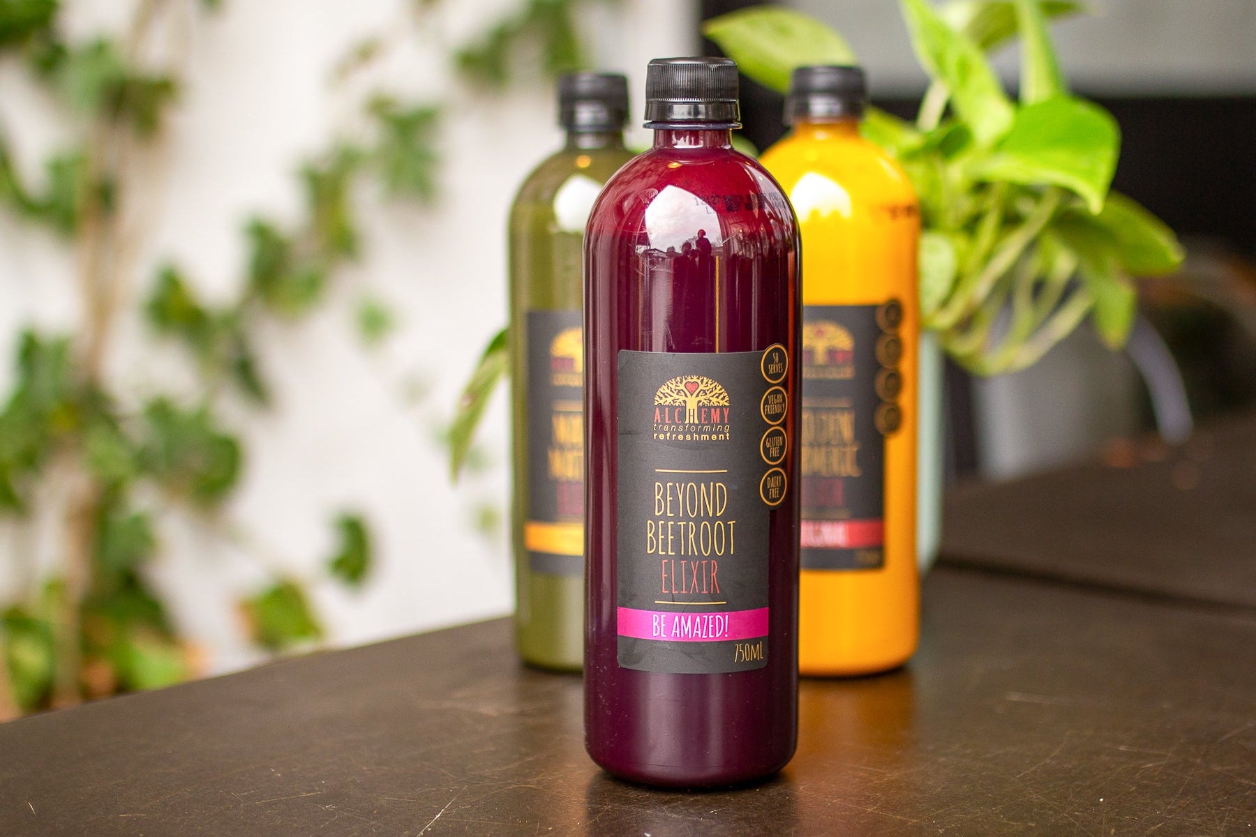 Three bottles of 'Beyond Beetroot Elixir' on a table with a blurred green plant background