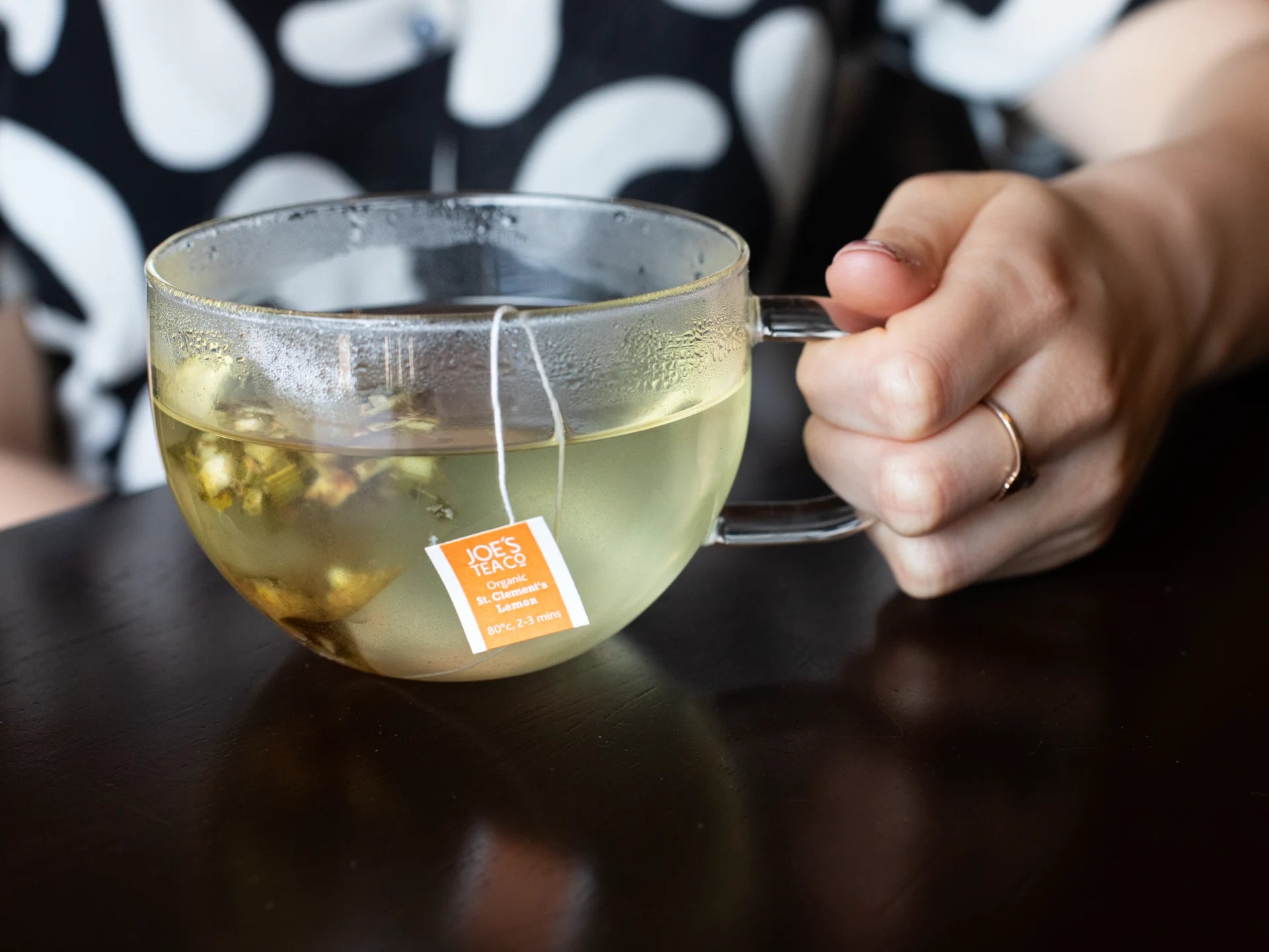 Hand holding a glass mug of tea with a tea bag, on a dark surface.