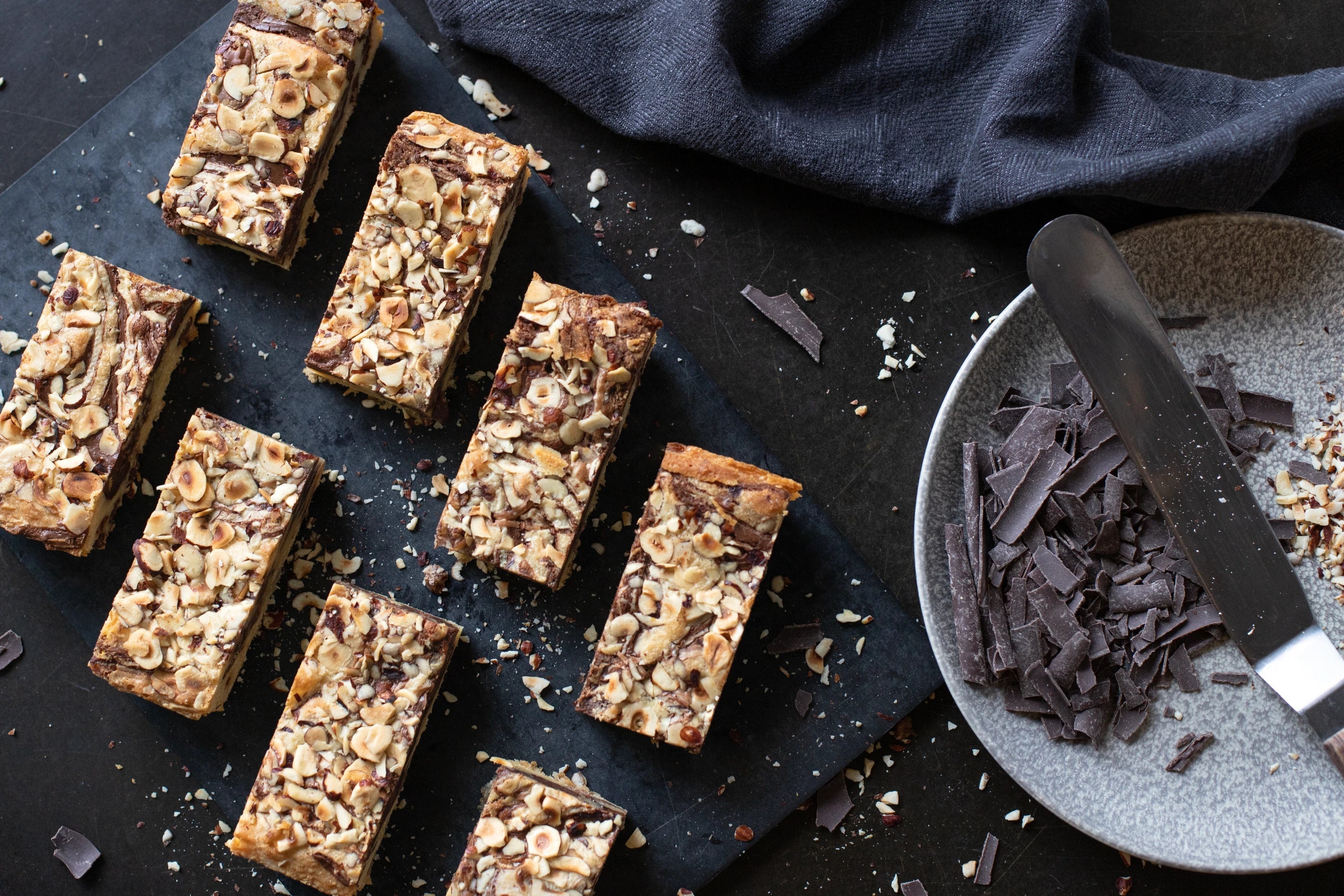 Hazelnut Blondie Nuts and chocolate bars on a dark surface with a bowl of chocolate shavings.