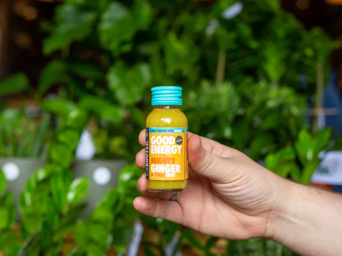 Hand holding a bottle of 'Unrooted Health Shot: Mighty Ginger' against a blurred green plant background