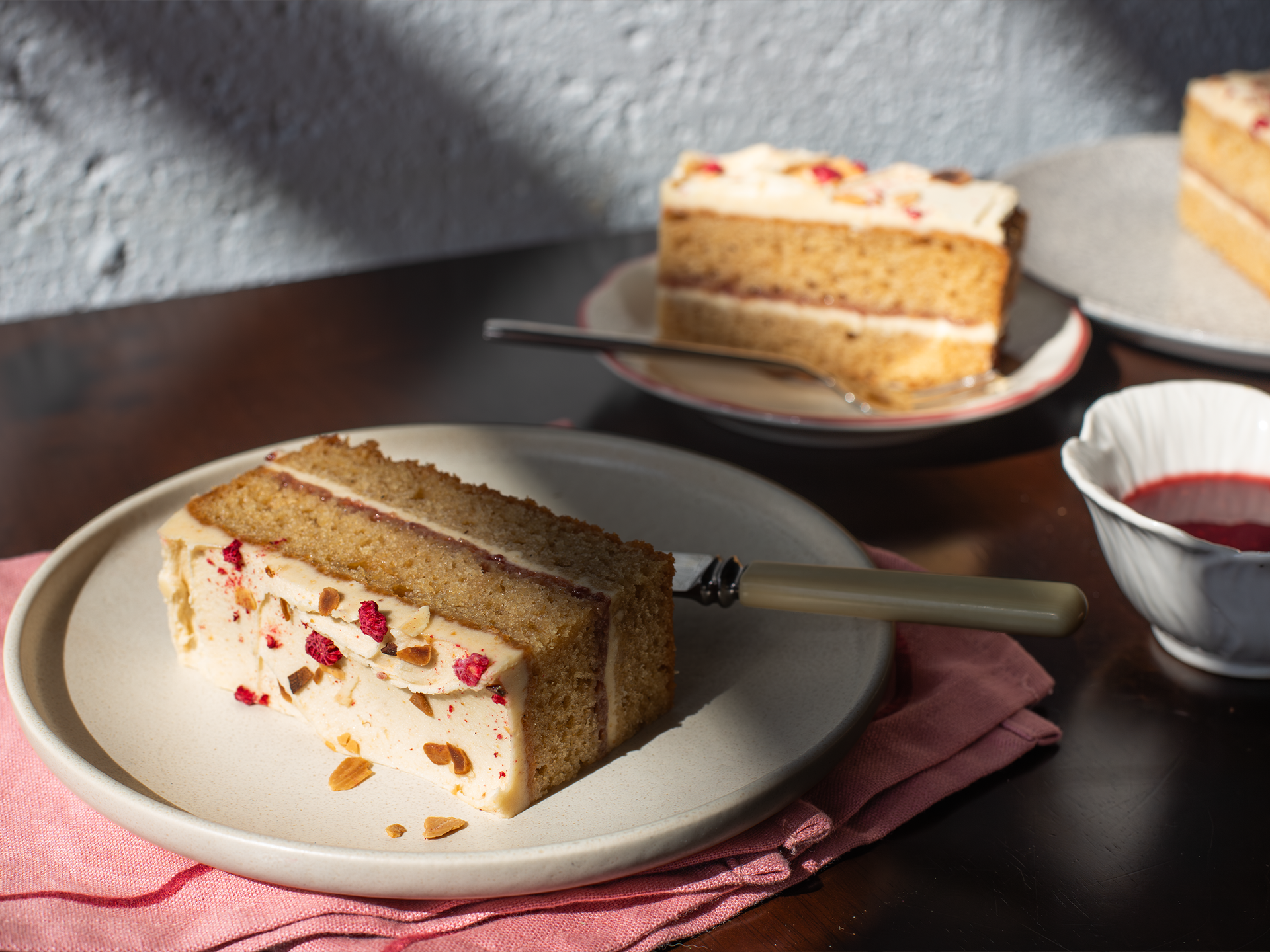 Two slices of Almond Butter and Jelly cake on plates with a pink napkin and a small bowl of red sauce in the background.