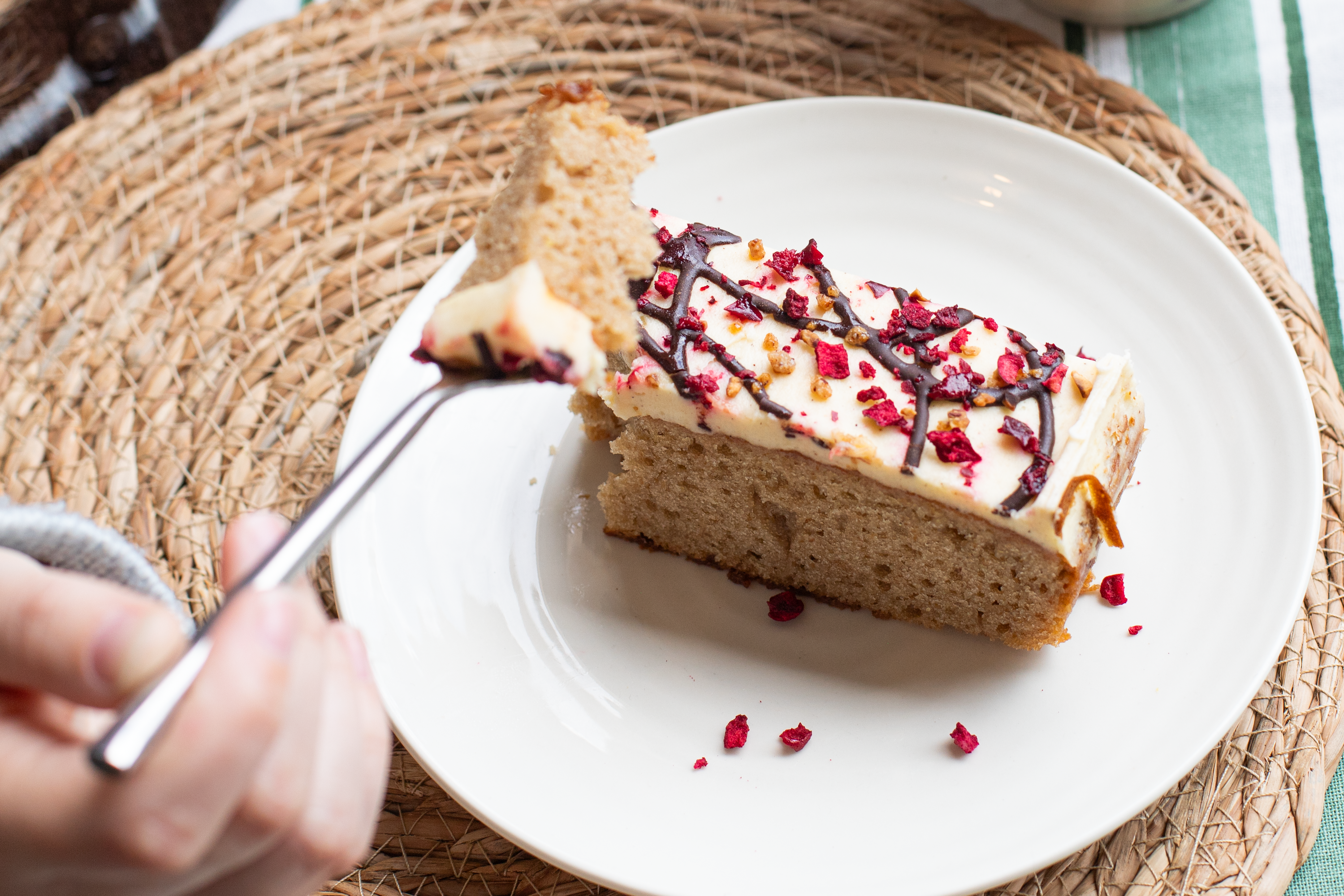 Slice of Banana Split cake with chocolate drizzle and red berries on a white plate, held by a hand with a fork.