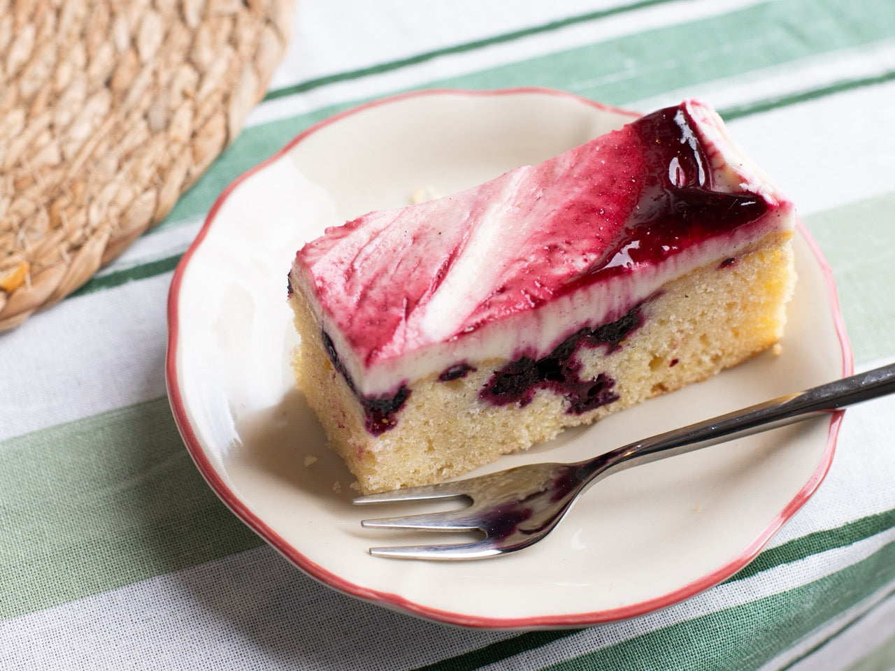 Slice of Blackcurrant Ripple cake with red fruit topping on a white plate with a fork, on a striped tablecloth.