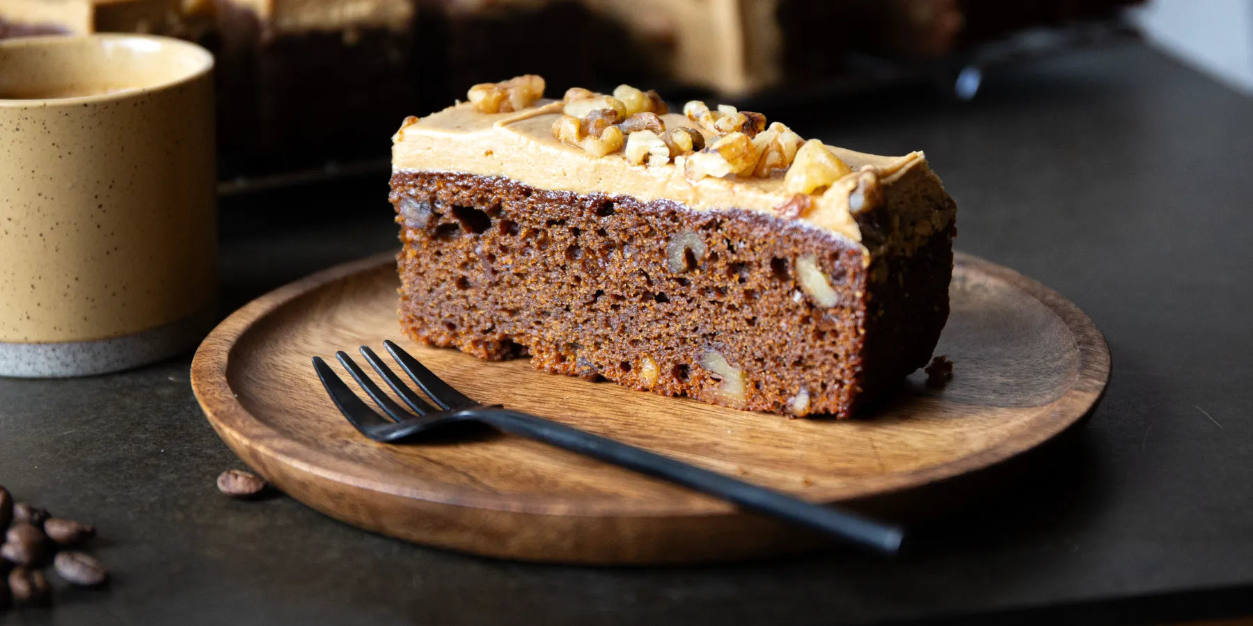 Slice of cake with frosting and nuts on a wooden plate with a fork, on a dark surface.