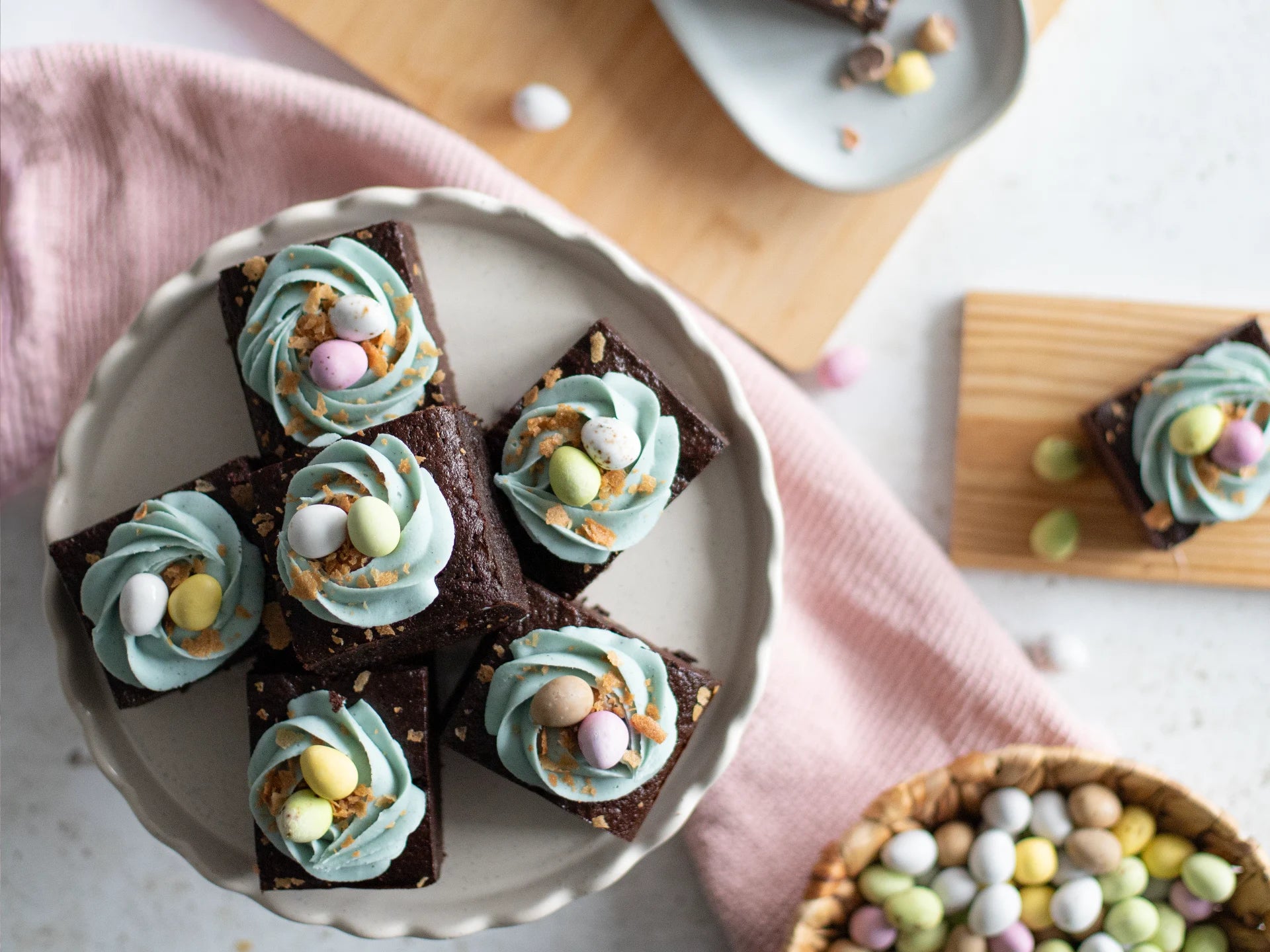 Chocolate Easter Nest Cake with blue frosting and colourful eggs on a white plate, with a pink cloth and additional candies in the background.