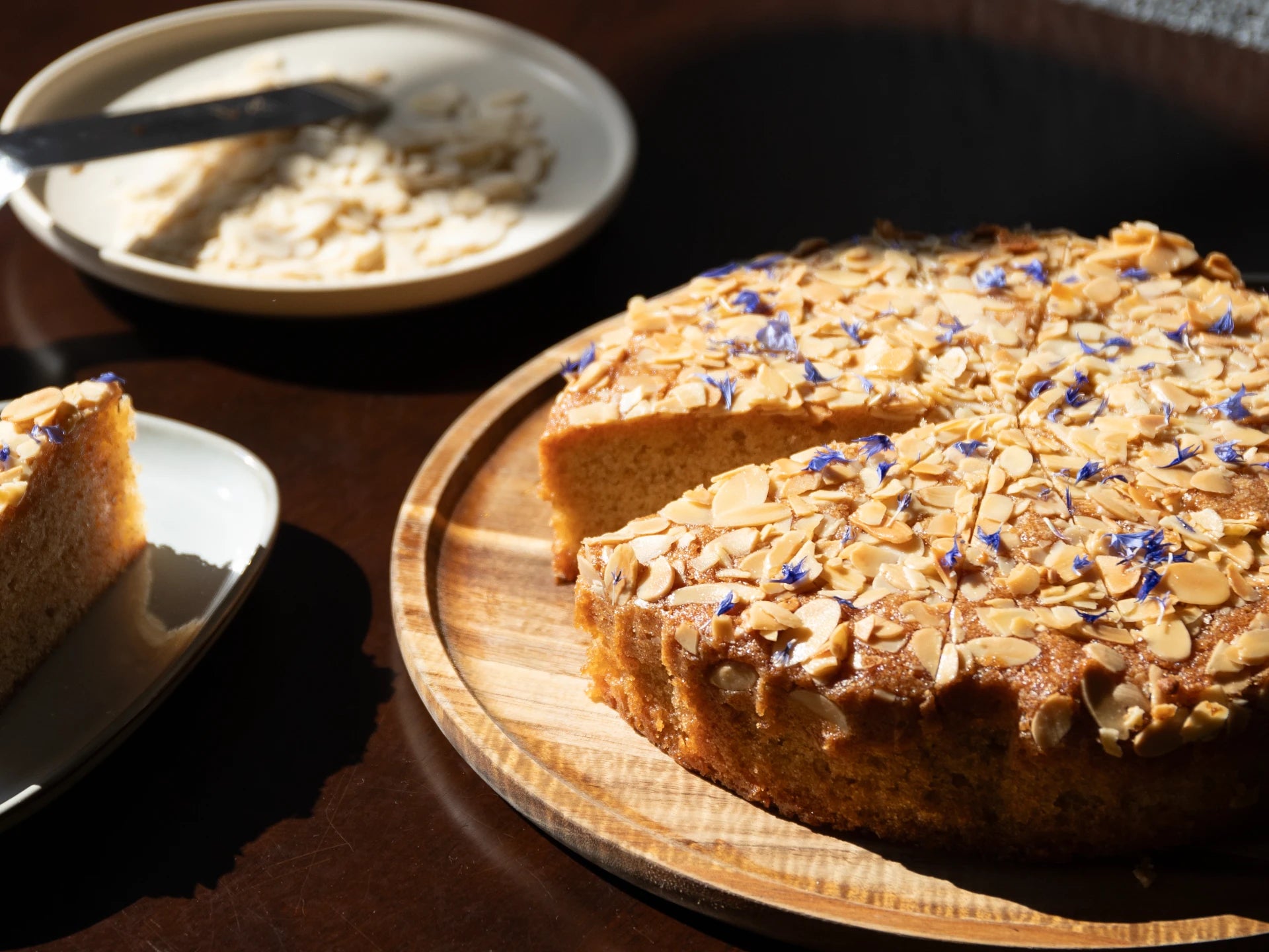 Honey Lavender Almond Round Cake with almonds and blueberries on a wooden board, with a slice taken out, on a dark surface.