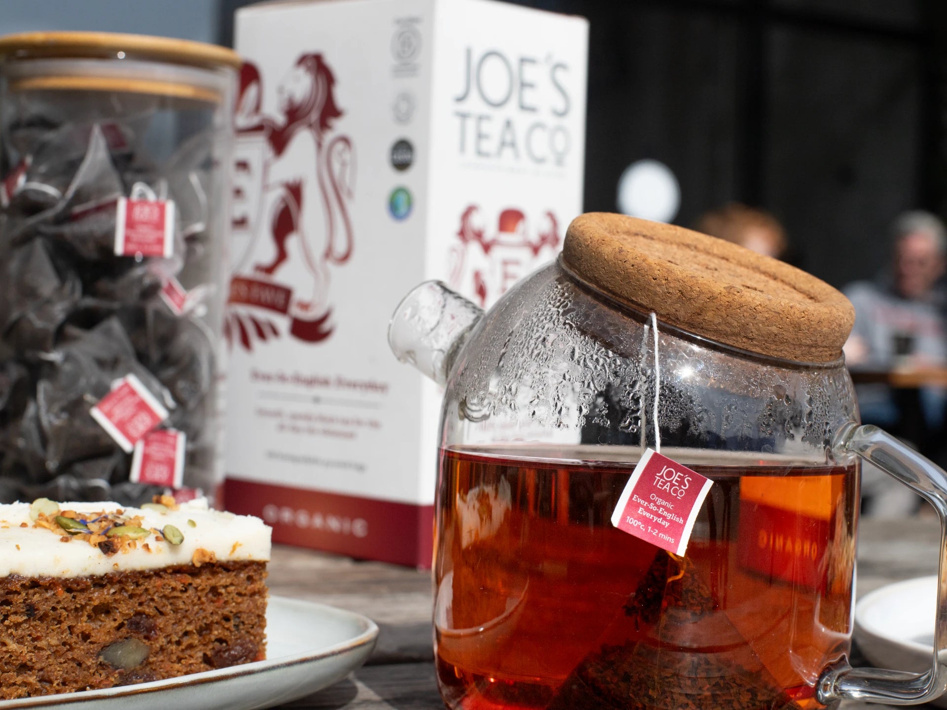 Glass teapot with Joe's Tea Co - Ever-So-English Breakfast tea and a cake on a table with 'Joe's Tea Co' branding.