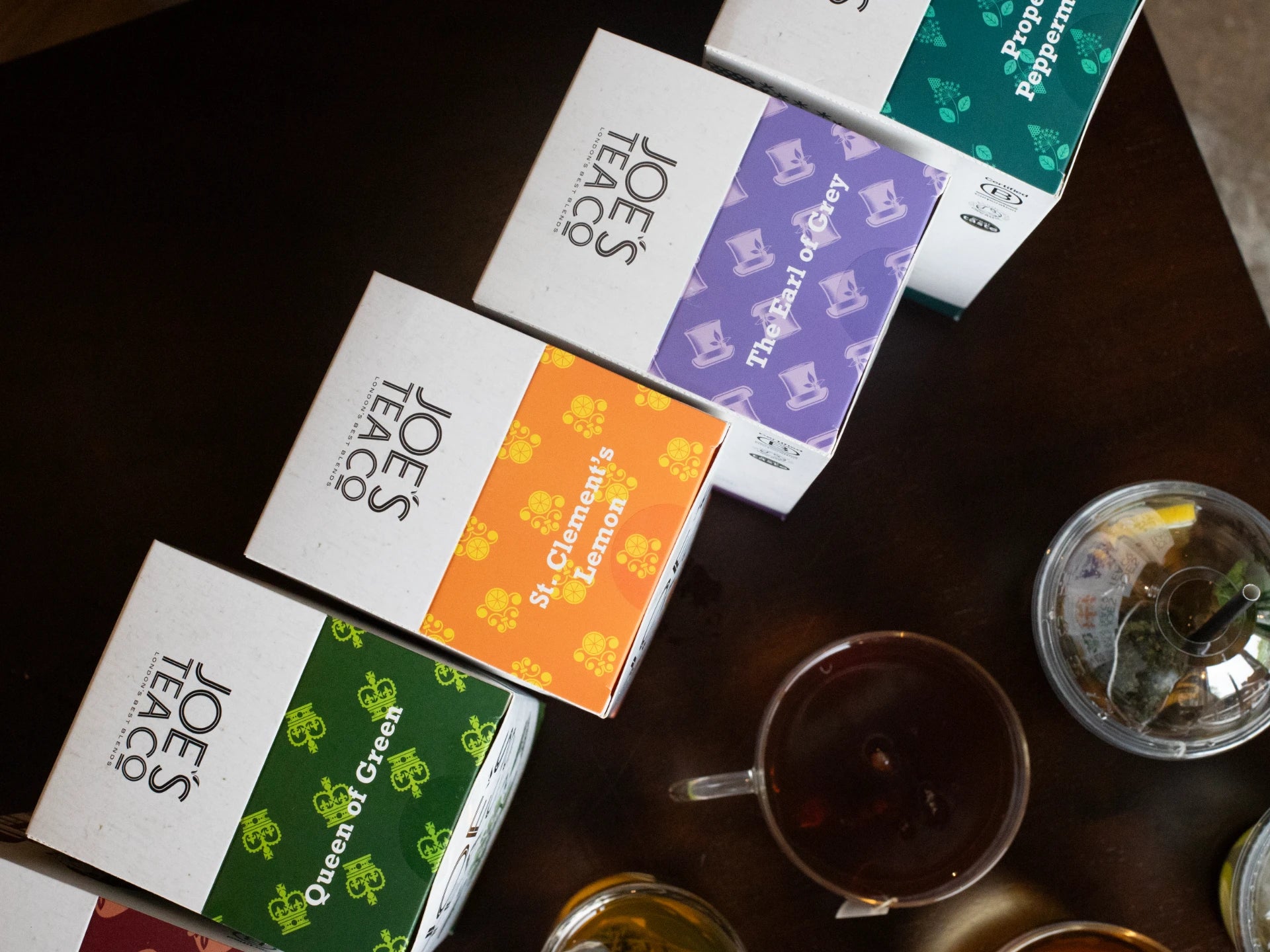 Assorted boxes of Joe's Teas Co. on a wooden surface with a tea cup and glass.