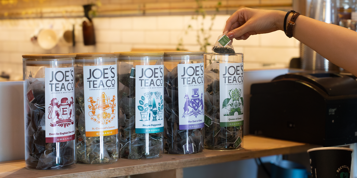Row of Joe's Tea Co. tea canisters on a wooden surface with a person opening one.