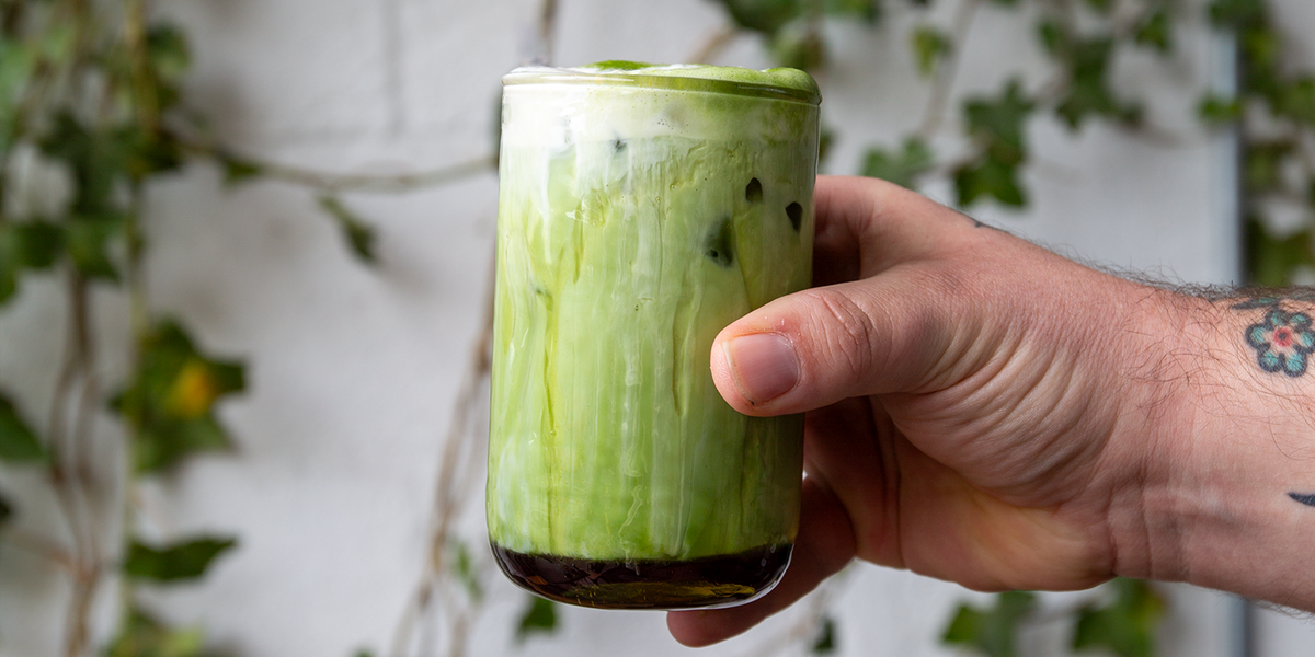 Hand holding a green Lav Honey Matcha iced drink in a clear glass against a blurred natural background