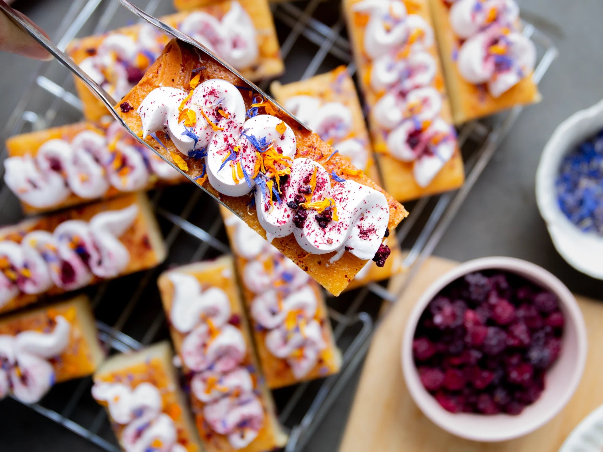 Decorative Lemon Blackberry Elderflower cake with white cream and colorful toppings on a cooling rack, with a bowl of berries in the foreground.