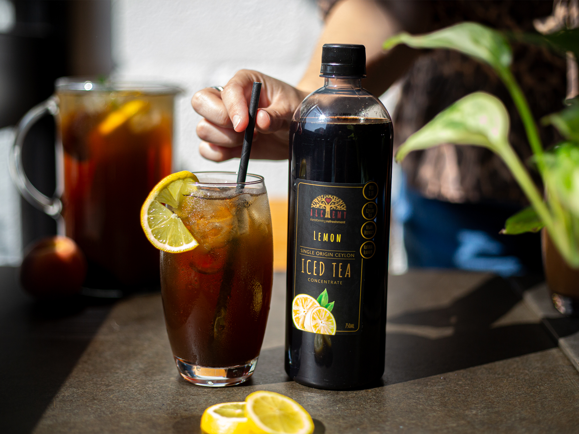 Bottle of Alchemy lemon iced tea with a glass of iced tea and lemon slices on a table.