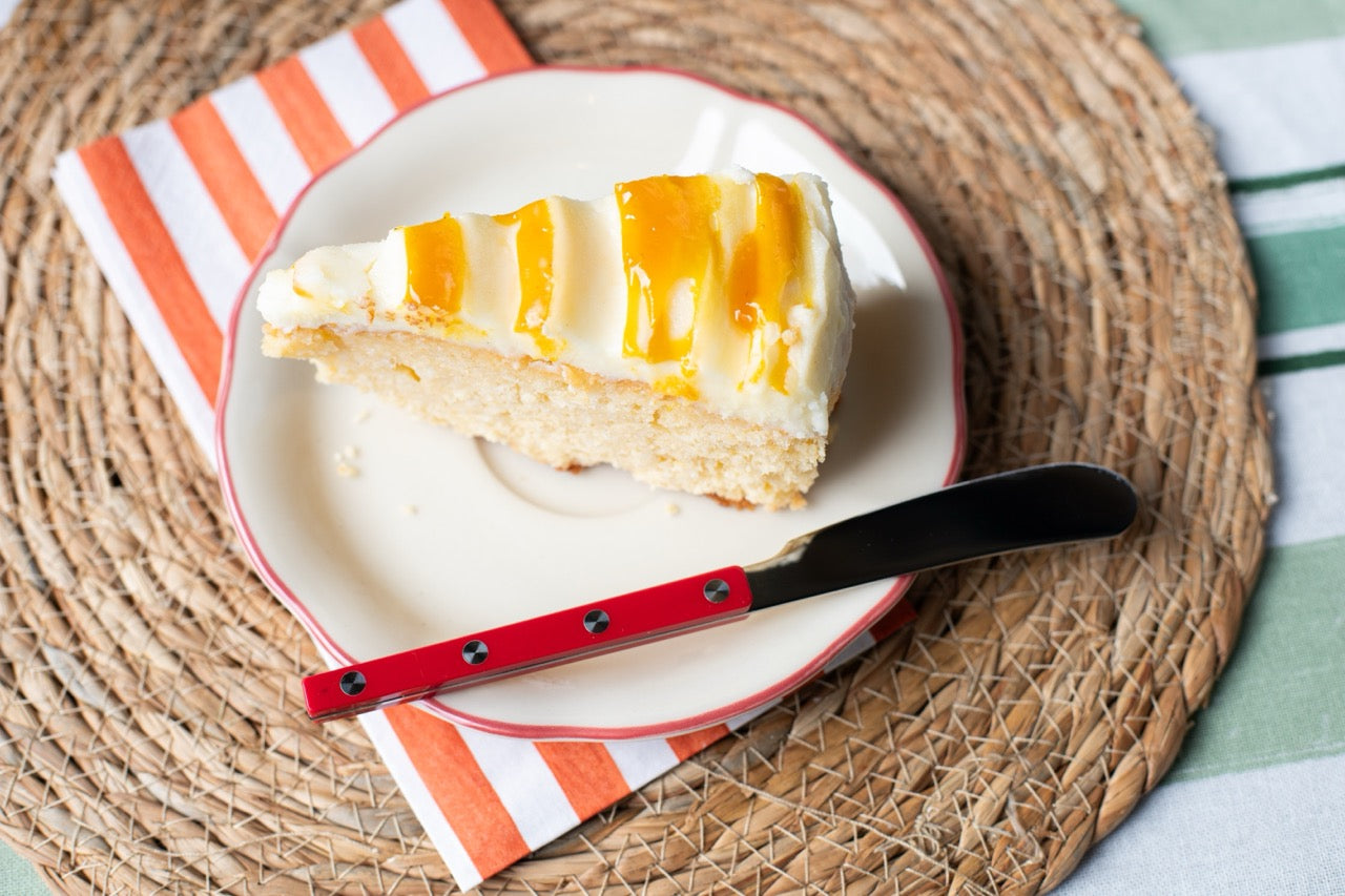 Slice of Mango Pineapple Coconut cake on a plate with a knife, placed on a woven mat with a striped cloth background.