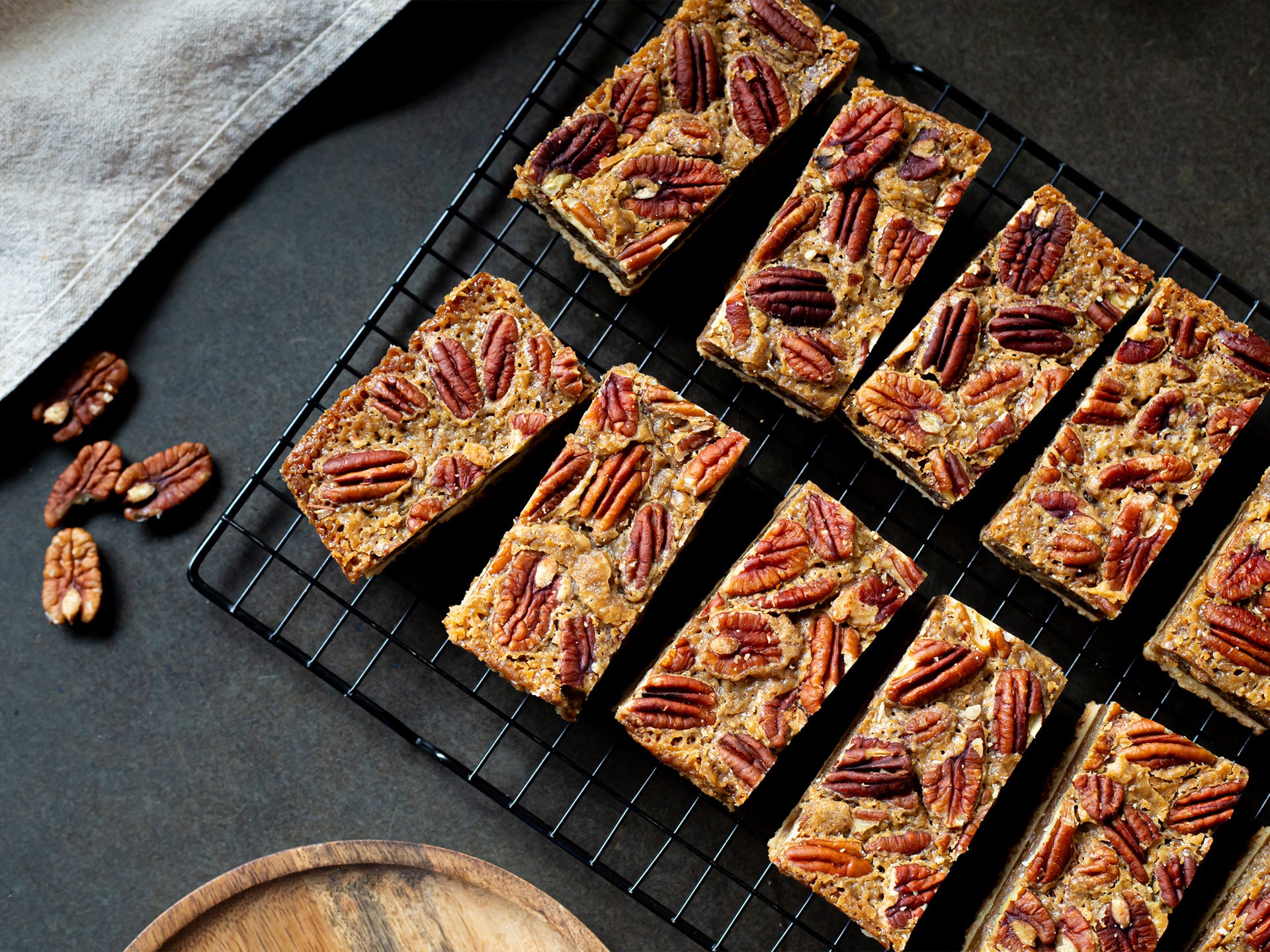 Pecan pie bars on a cooling rack with scattered pecans on a dark surface.