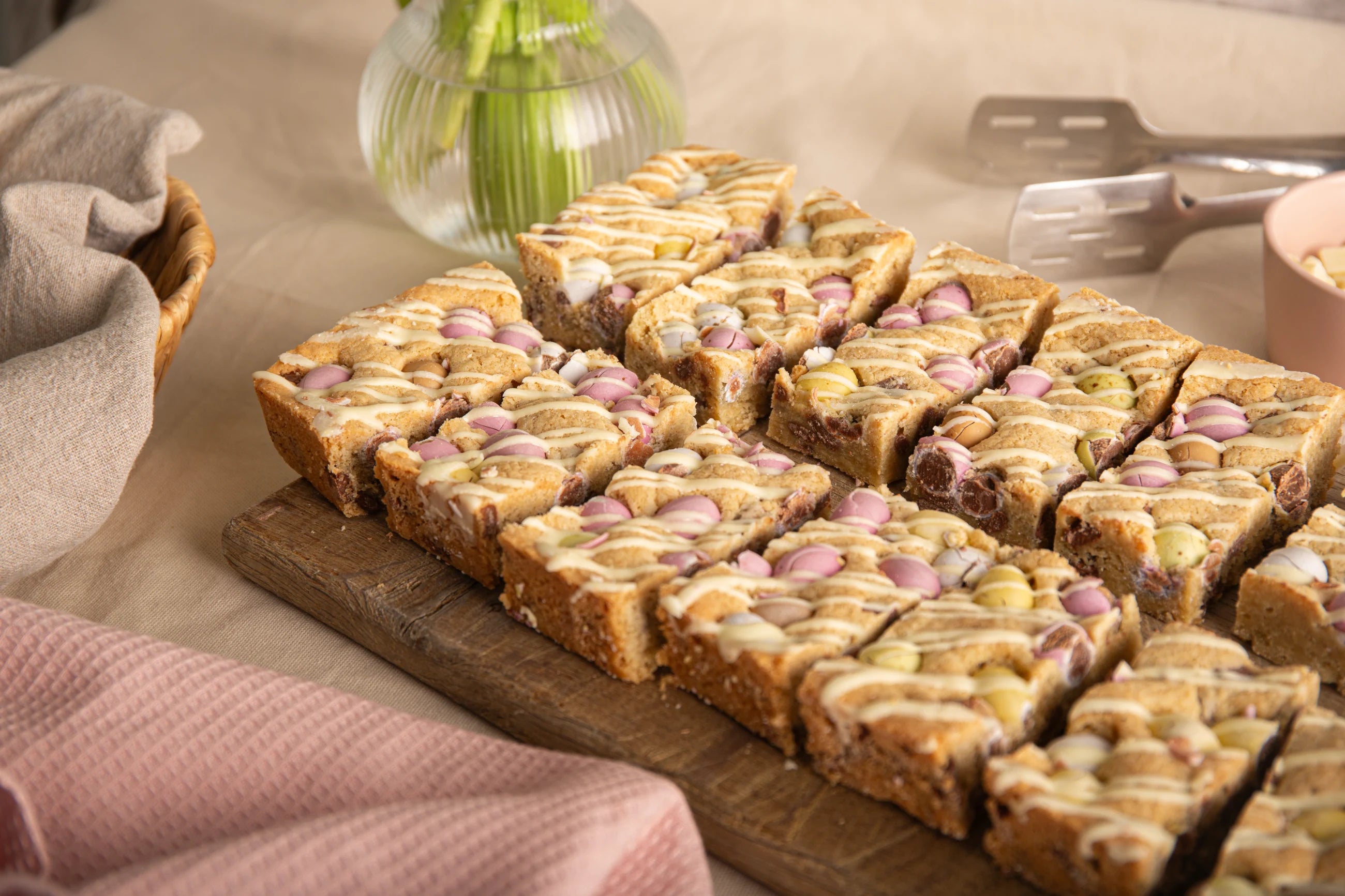 Sliced Mini Egg Cookie Bars with candy pieces on a wooden board, with a pink towel and vase in the background.