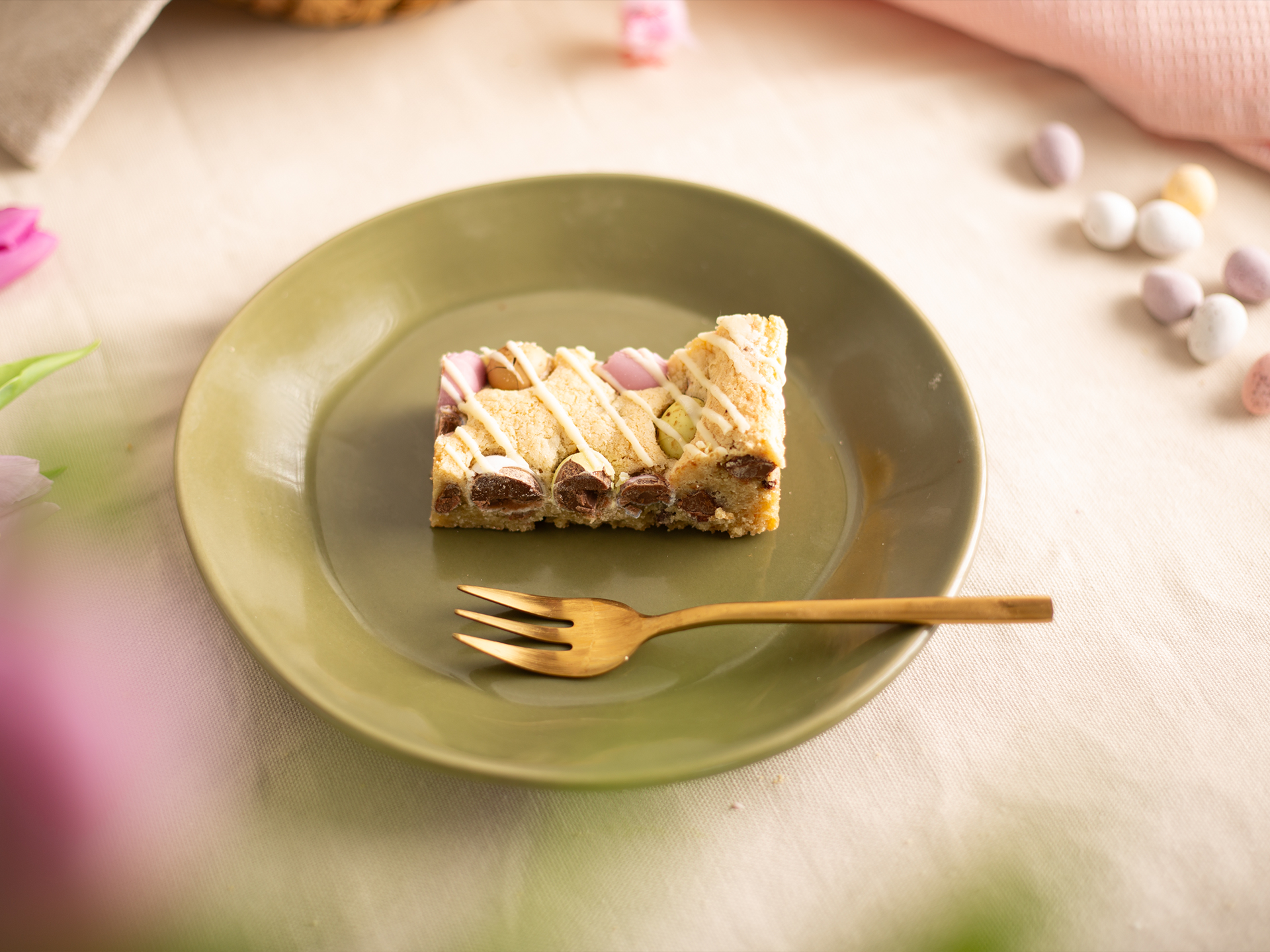 Slice of Mini Egg Cookie Bar on a green plate with a gold fork, surrounded by colorful Easter eggs on a light background.