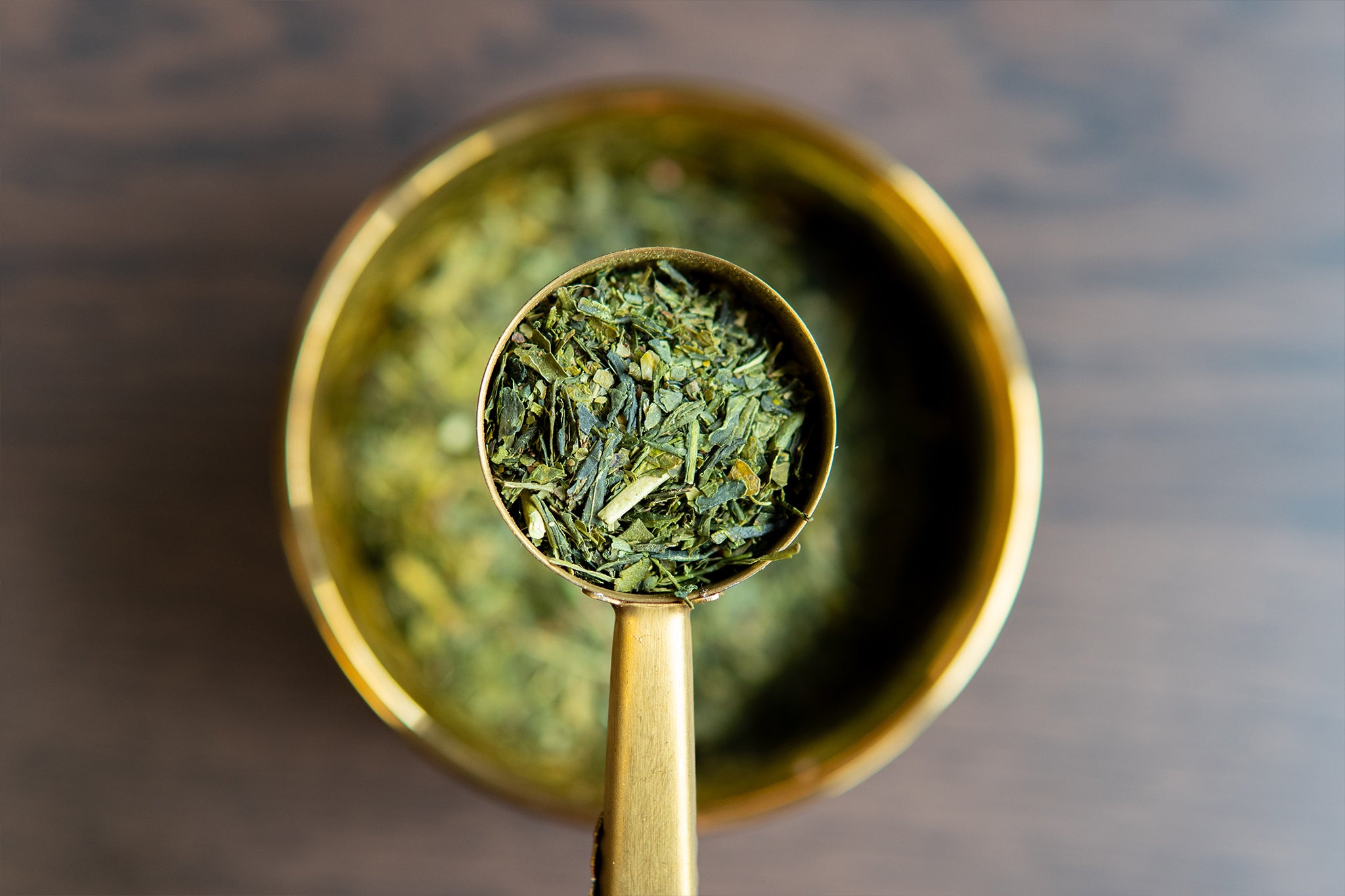 A spoonful of loose leaf Sencha green tea placed in a brass bowl filled with the same tea.