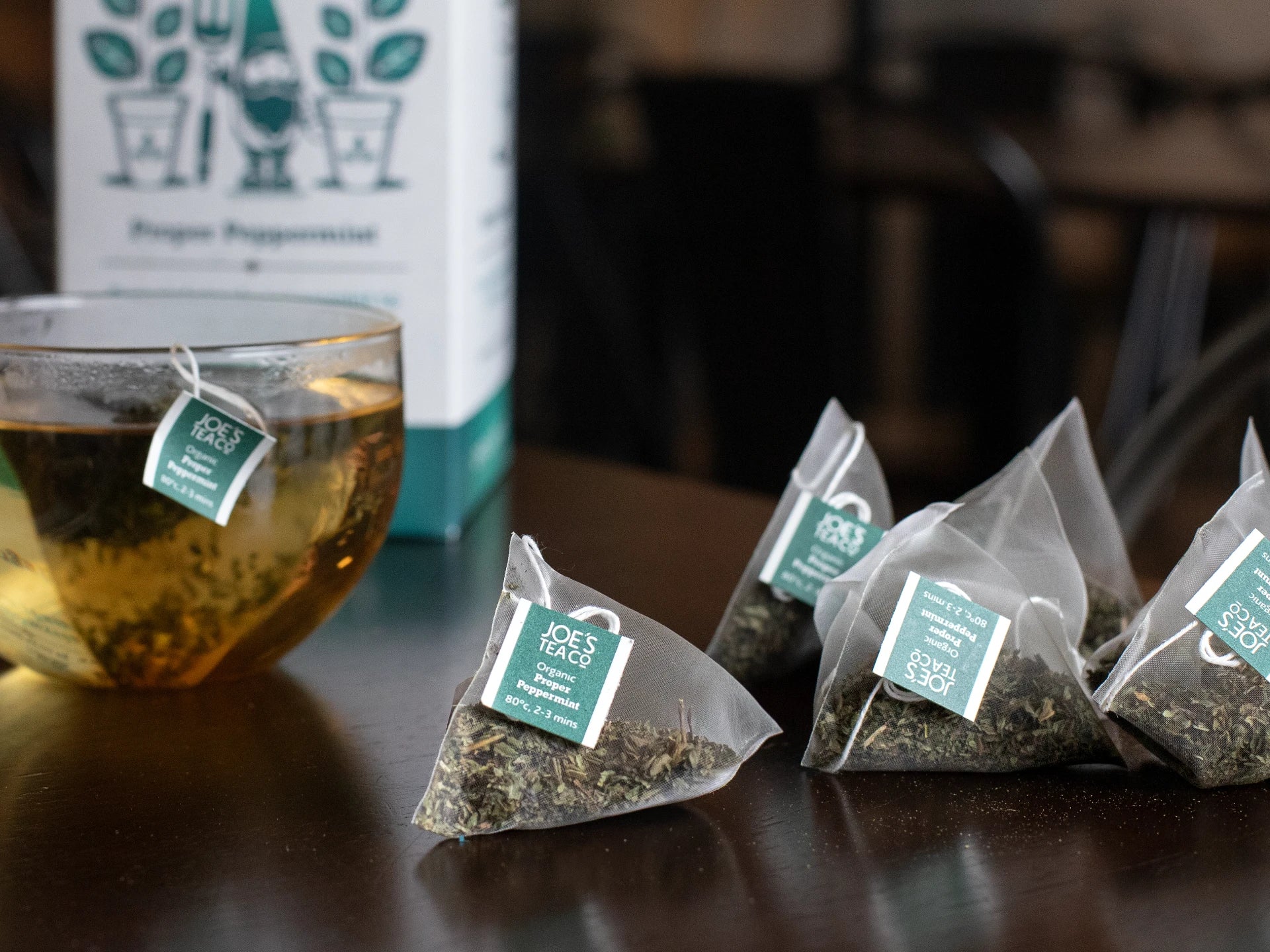 Joe's Tea Co - Proper Peppermint Tea bags with green labels on a wooden surface, with a glass of tea and packaging in the background.