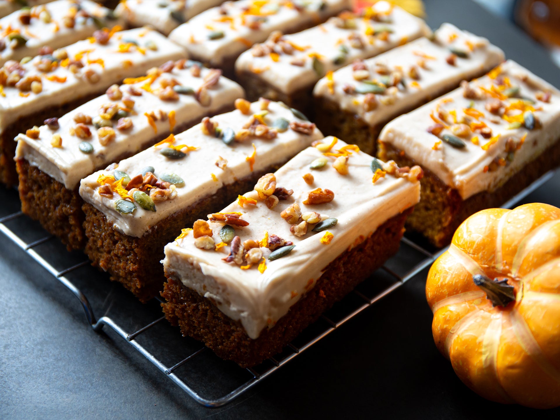 Slices of pumpkin and maple cake with white frosting and pumpkin seeds on a cooling rack, with a small pumpkin beside them.