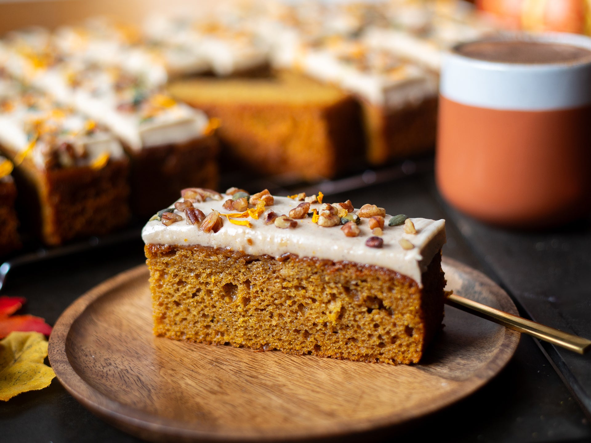 A Slice of pumpkin cake with cream cheese frosting on a wooden plate, served with a hot chocolate