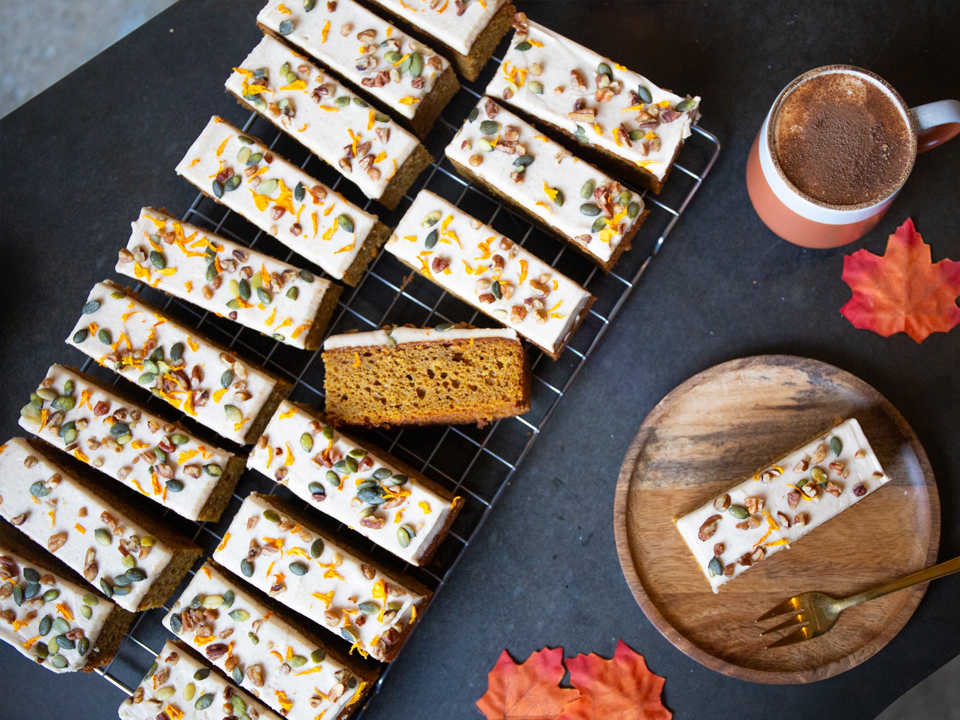 Pumpkin cake slices with cream cheese frosting and autumn seeds, accompanied by a cup of coffee and decorative leaves.