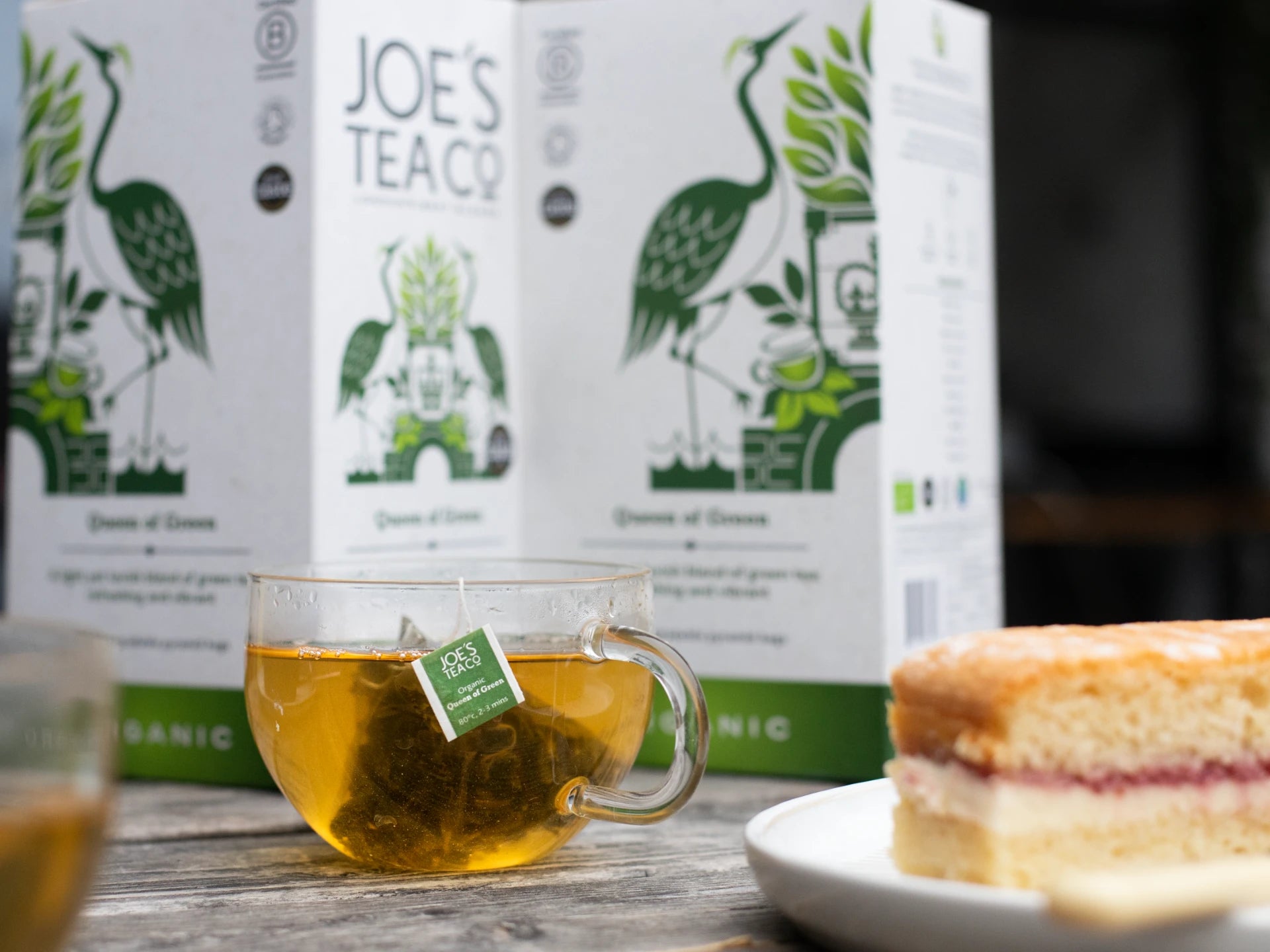 Tea cup with green tea and a cake on a wooden table with 'Joe's Tea Co.' boxes in the background.