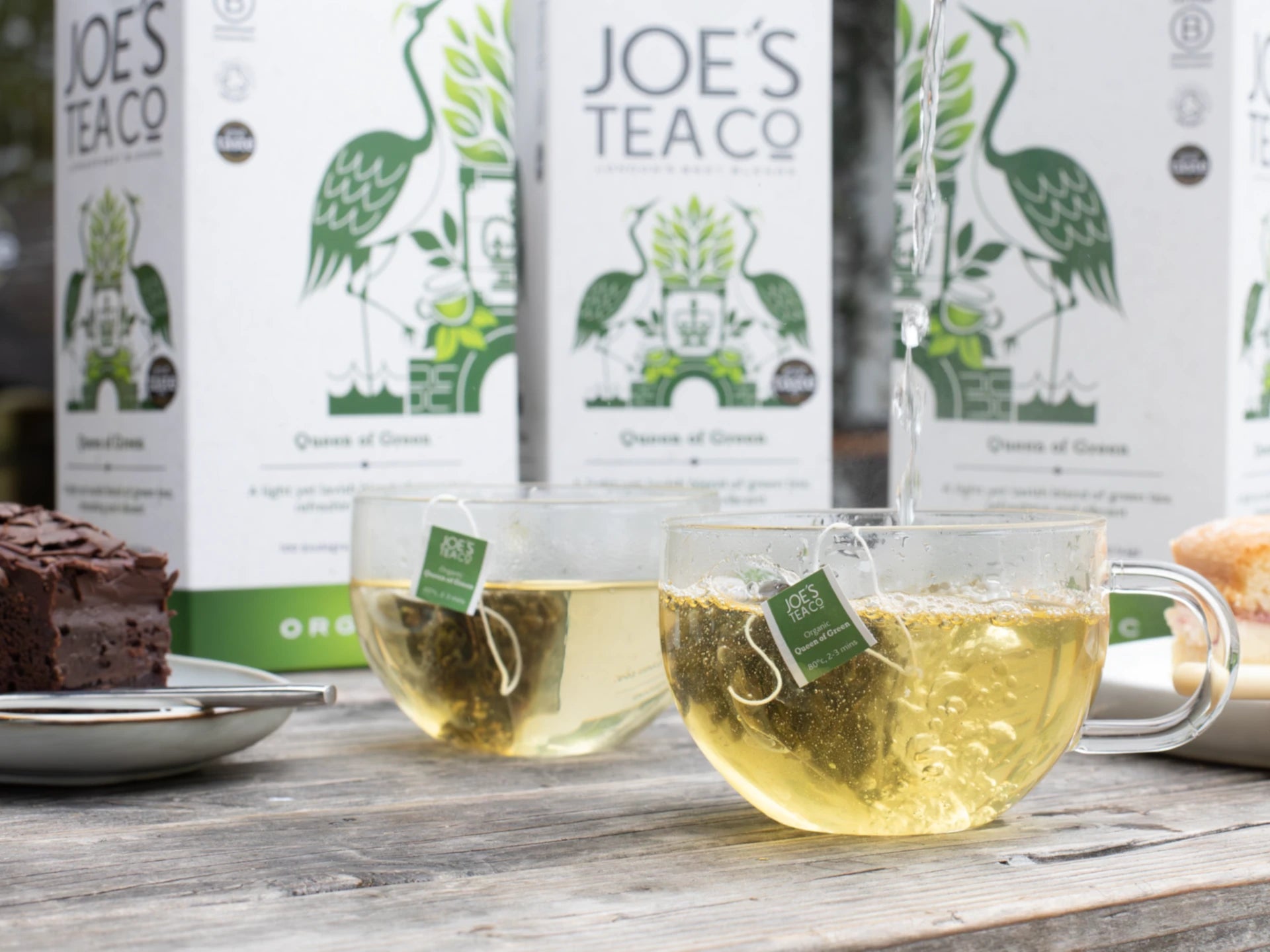 Two cups of tea with Joe's Tea Co. bags on a wooden table with tea boxes in the background.