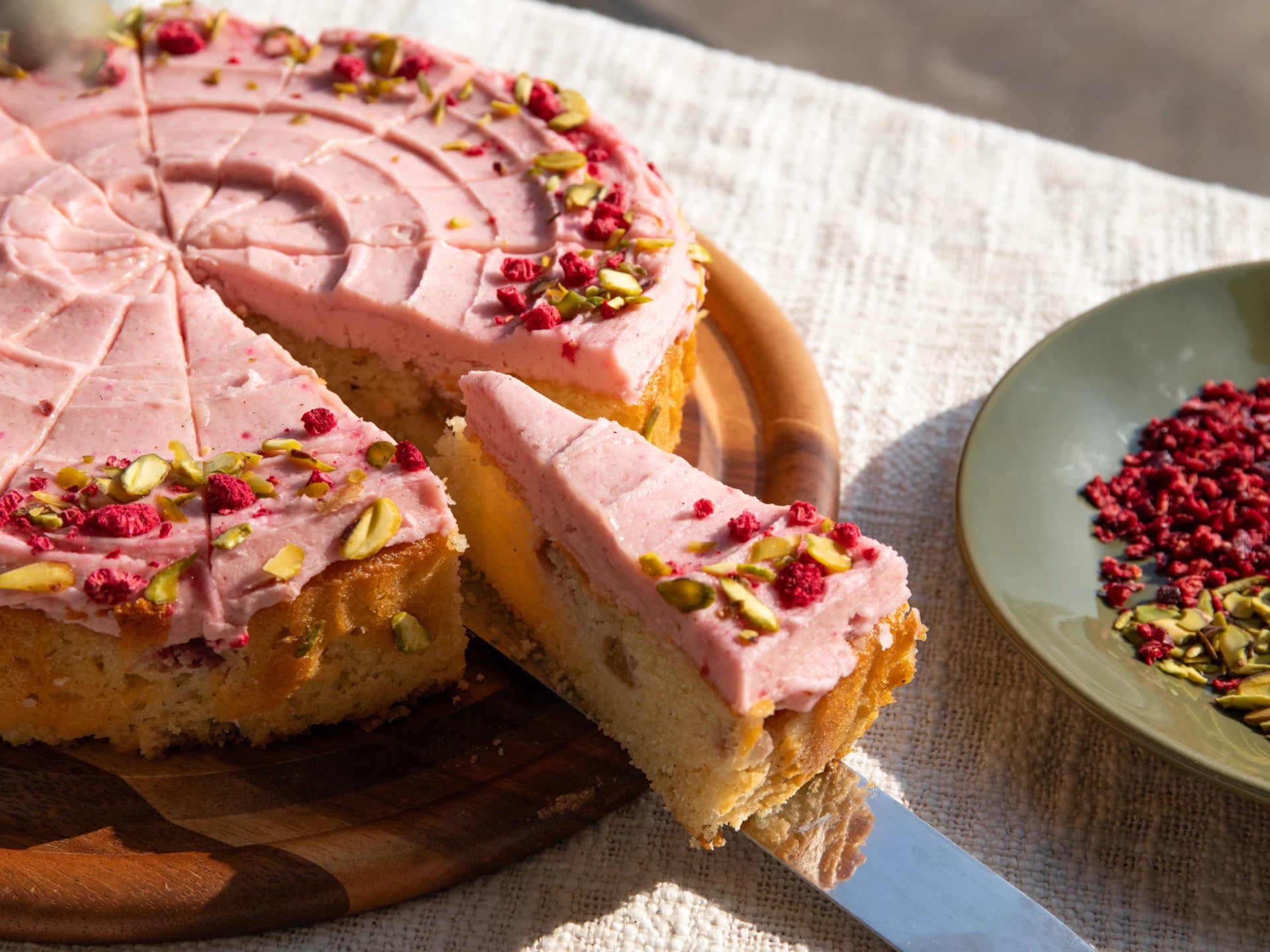 Pink frosted Raspberry Rhubarb Cake  with a slice removed on a wooden board, with a plate of red and green toppings in the background.