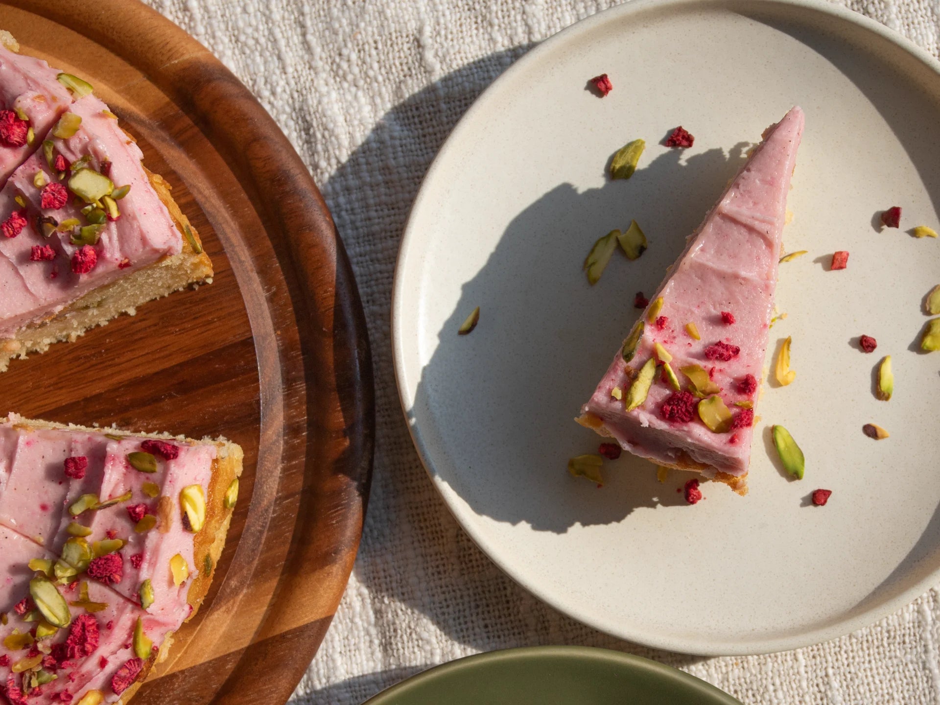 Slice of pink Raspberry Rhubarb Cake  with raspberry and pistachio on a white plate, with the rest of the cake on a wooden board.