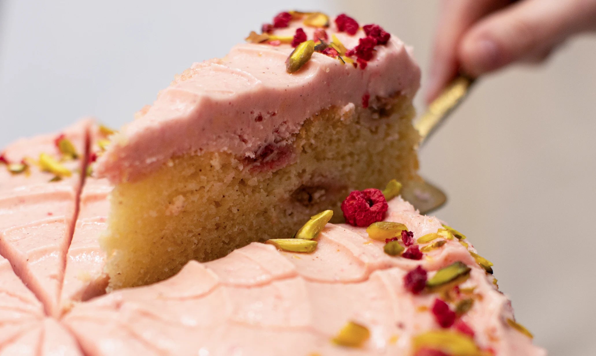 Slice of pink Raspberry & Rhubarb cake with raspberries and pistachios being lifted from the cake.