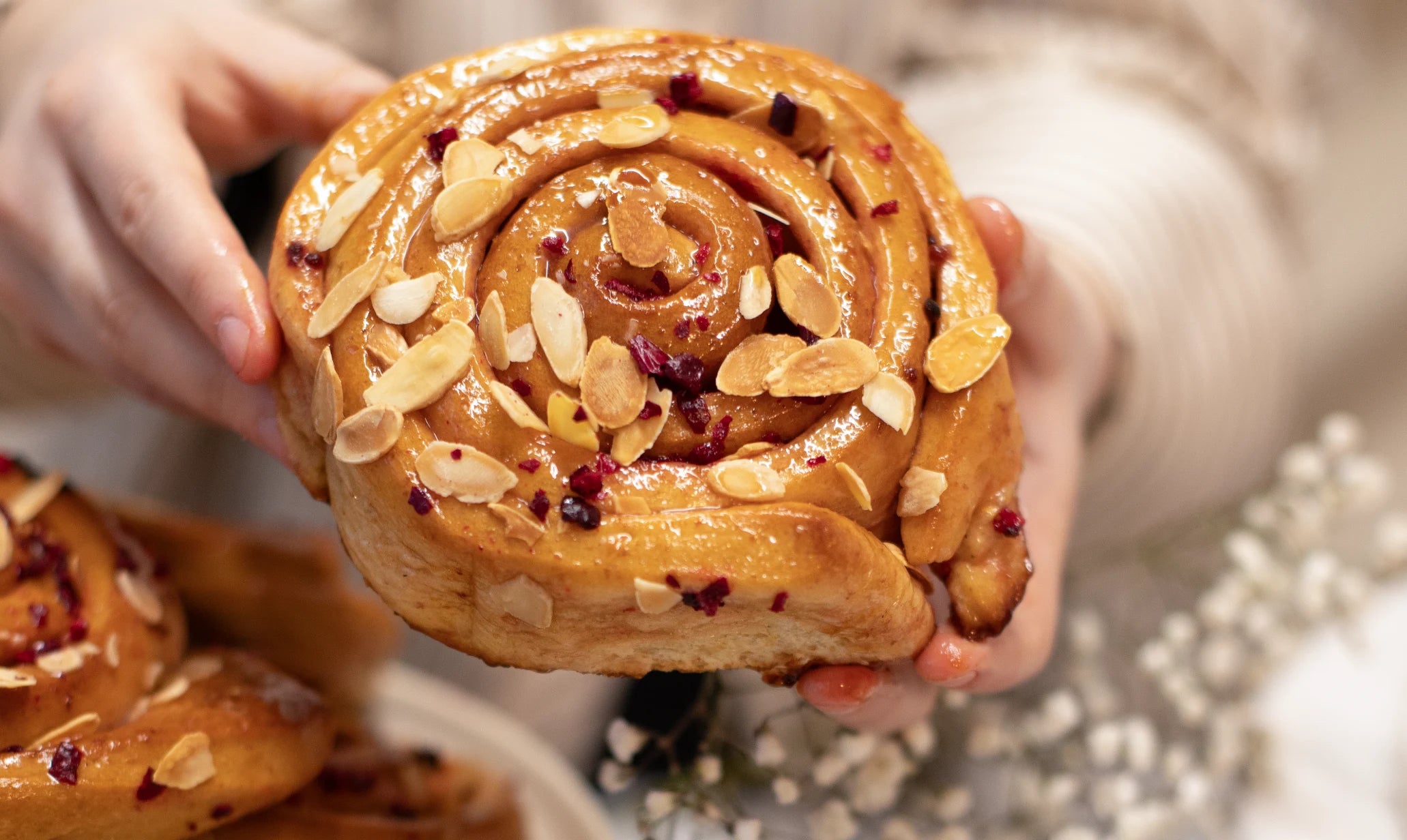 Hand holding a Sticky Cherry Bakewell Bun with almonds and seeds, blurred background