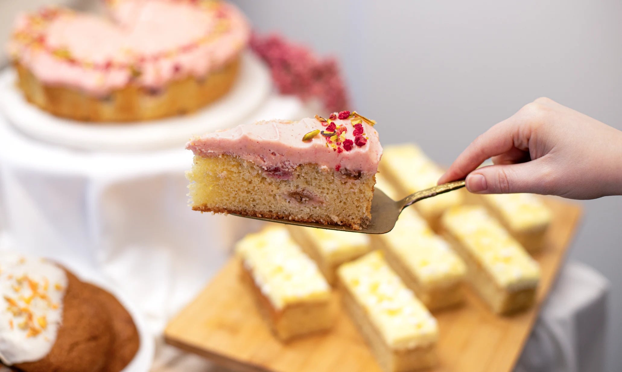 Slice of cake being lifted with a spatula, surrounded by other cakes on a wooden board.